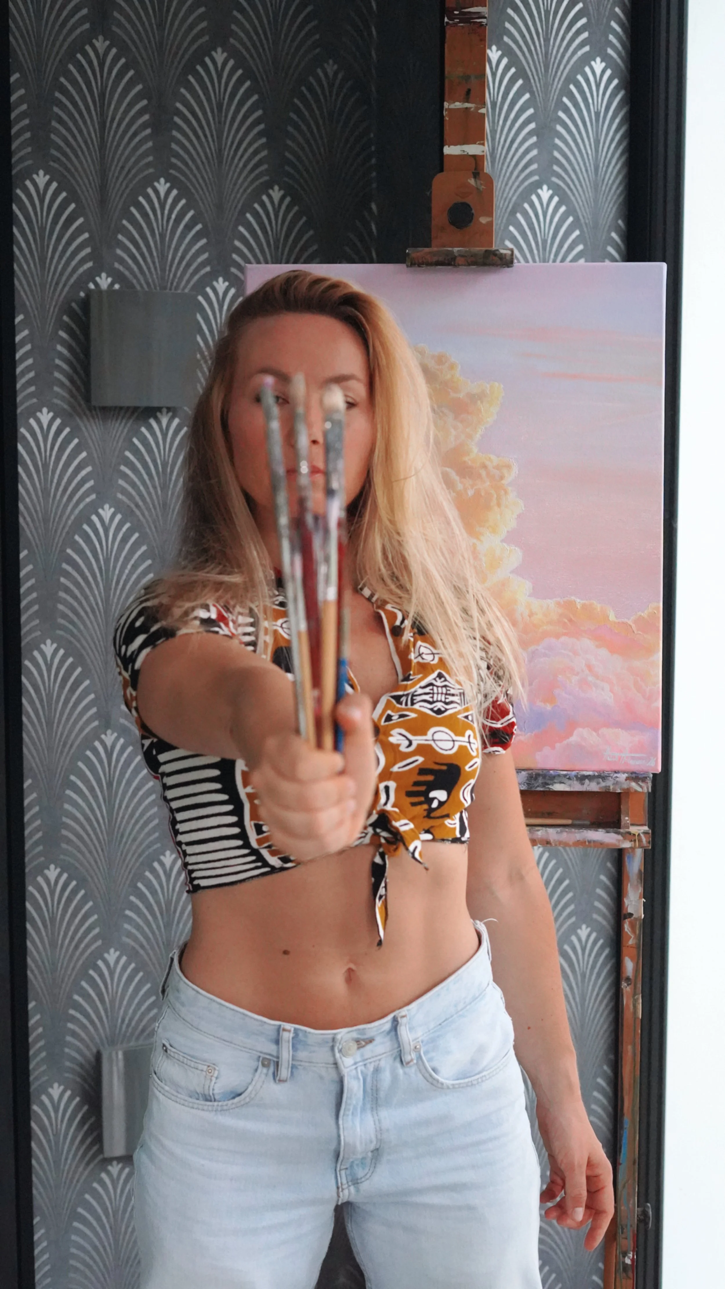 A woman with blonde hair in a patterned crop top and light jeans holding paintbrushes toward the camera inside an art studio with a decorative wallpaper and a painting of clouds and sky in the background.