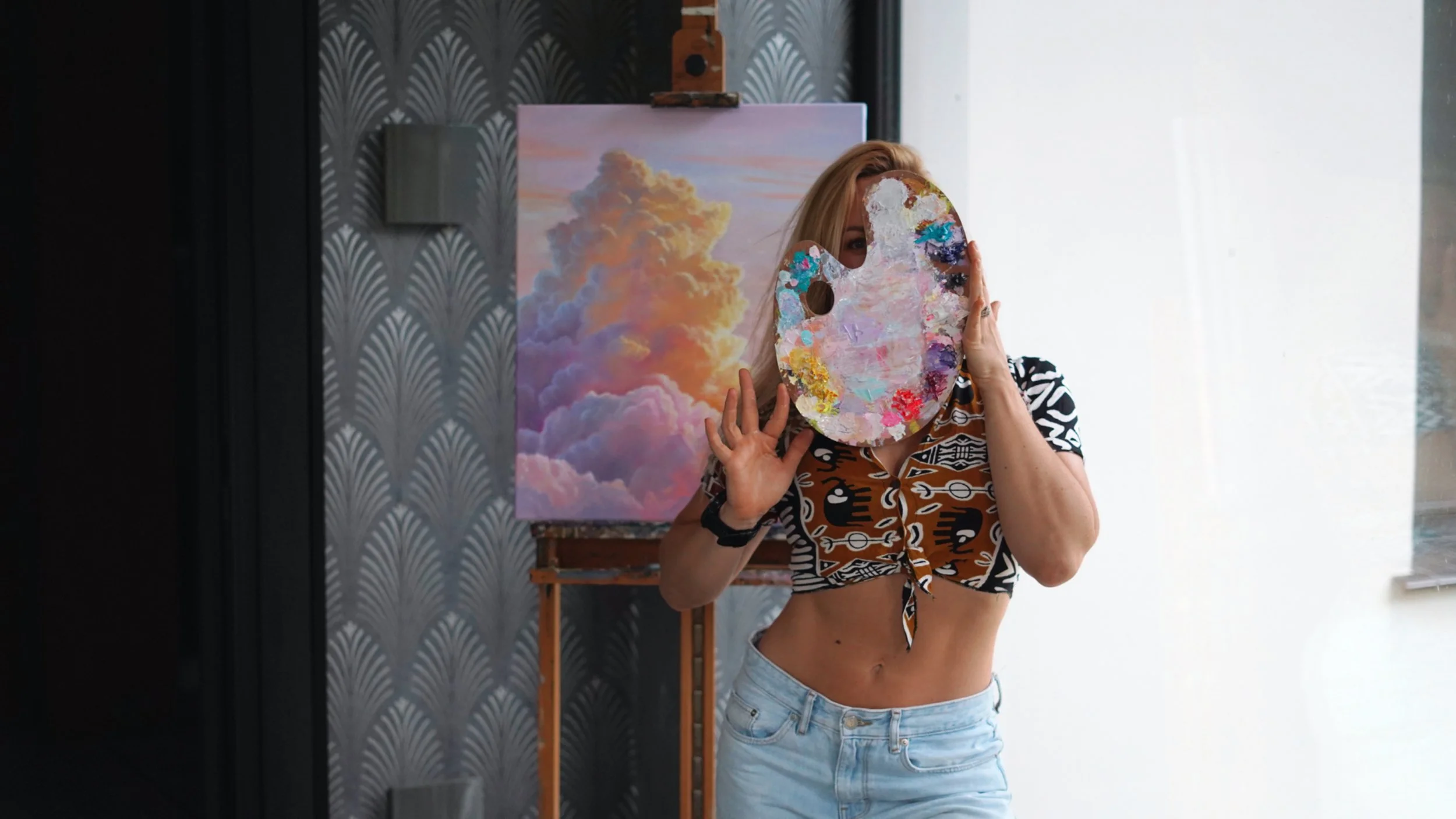 A woman with long blonde hair holding a painter's palette in front of her face, standing in front of a colorful cloud painting on an easel inside a gallery or studio with patterned wallpaper