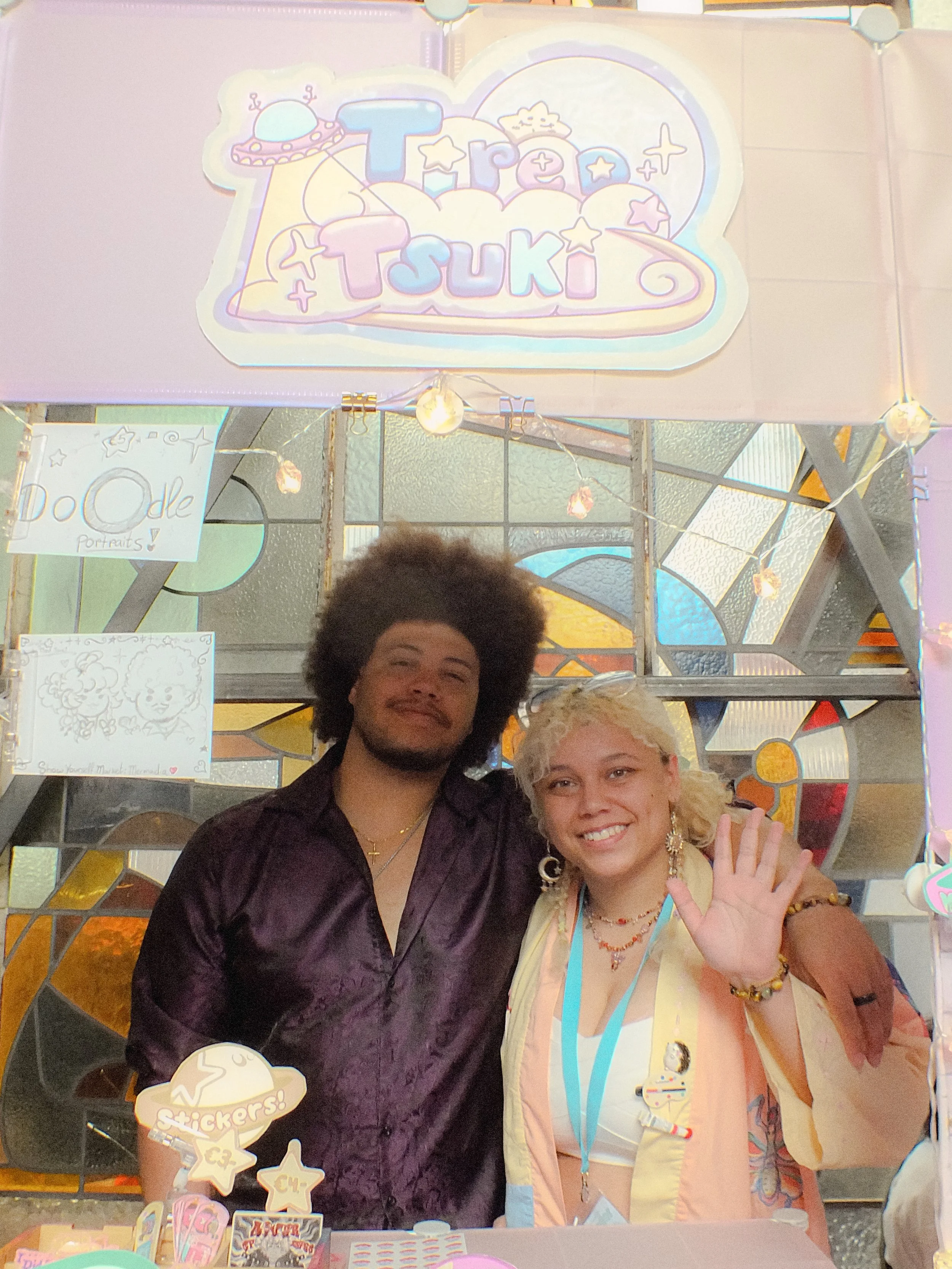 Two people, a man with dark curly hair and a woman with blonde hair, smiling and standing close together at a colorful event booth. The woman is waving and wearing jewelry. The booth has a sign saying 'Trea Tsuki' with toy and sticker displays, and decorative lights are in the background.