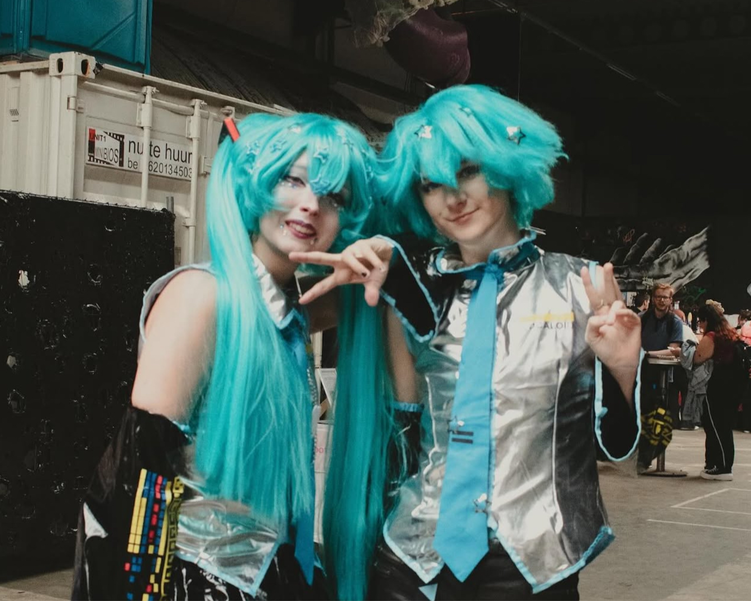 Two people dressed in vibrant blue wigs and costumes posing together indoors at a crowded event.