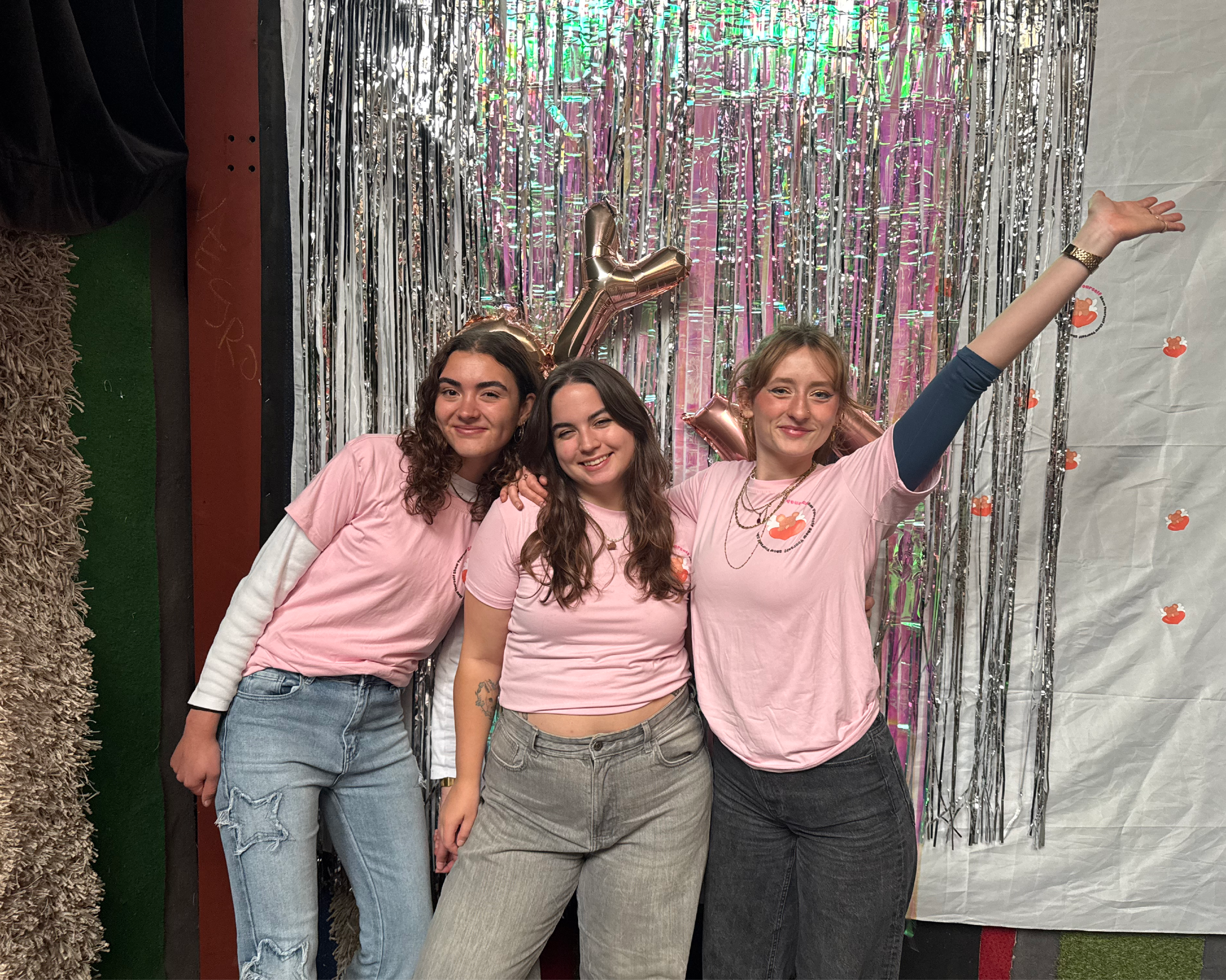 Three young women wearing pink T-shirts with a logo, standing close together in front of metallic and decorative backdrop, with one woman raising her arm and making a peace sign gesture
