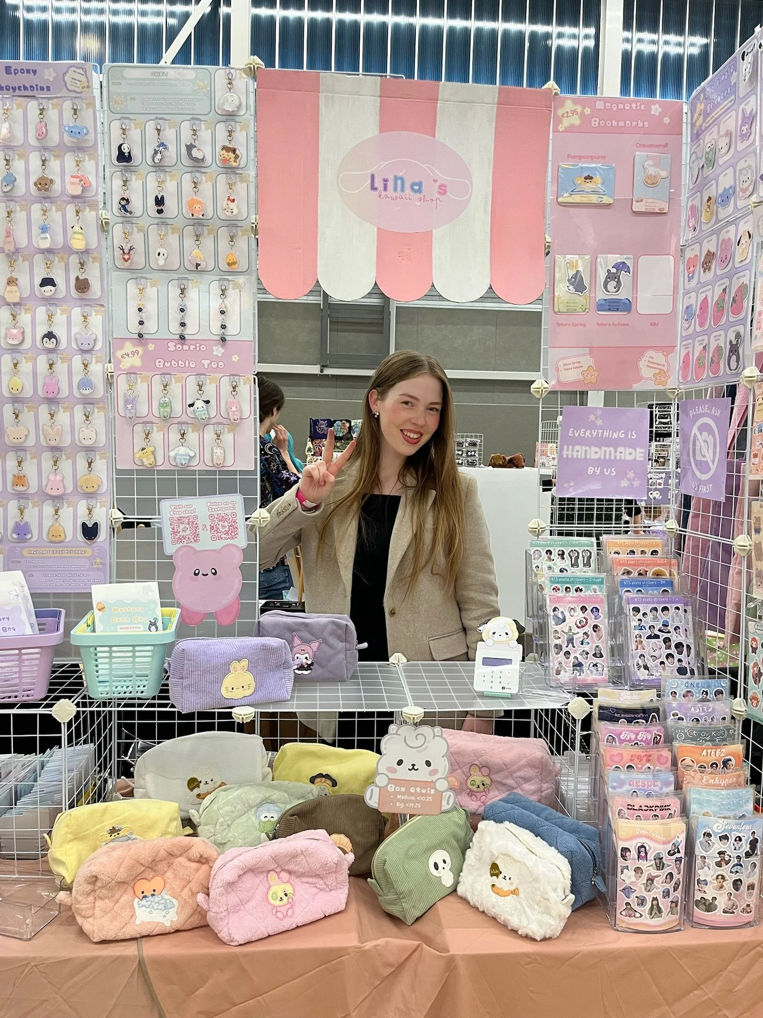 A woman smiling and making a peace sign behind a display table with various plush toys, bags, and accessories in pastel colors, at a booth labeled 'Lili's kawaii shop' with decorative signs and merchandise on several hooks and shelves.