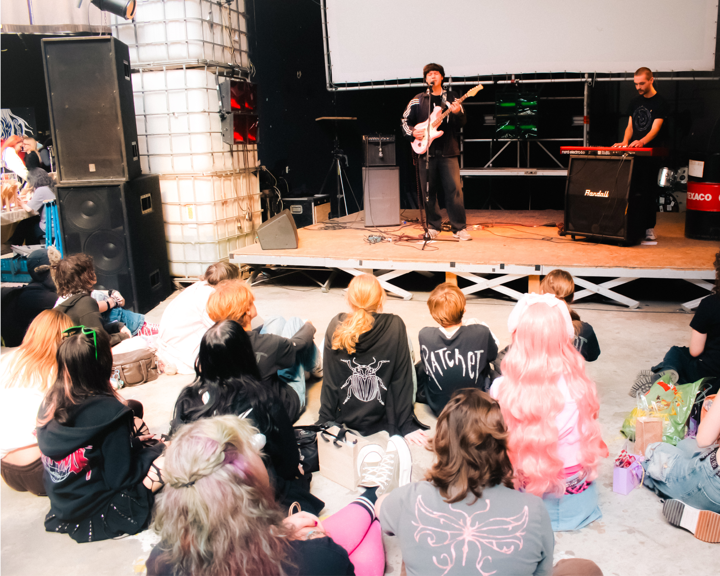 A musician performing on stage with a microphone, playing an electric guitar, while another person playing a keyboard stands nearby. An audience is seated on the floor in front of the stage, watching the performance.