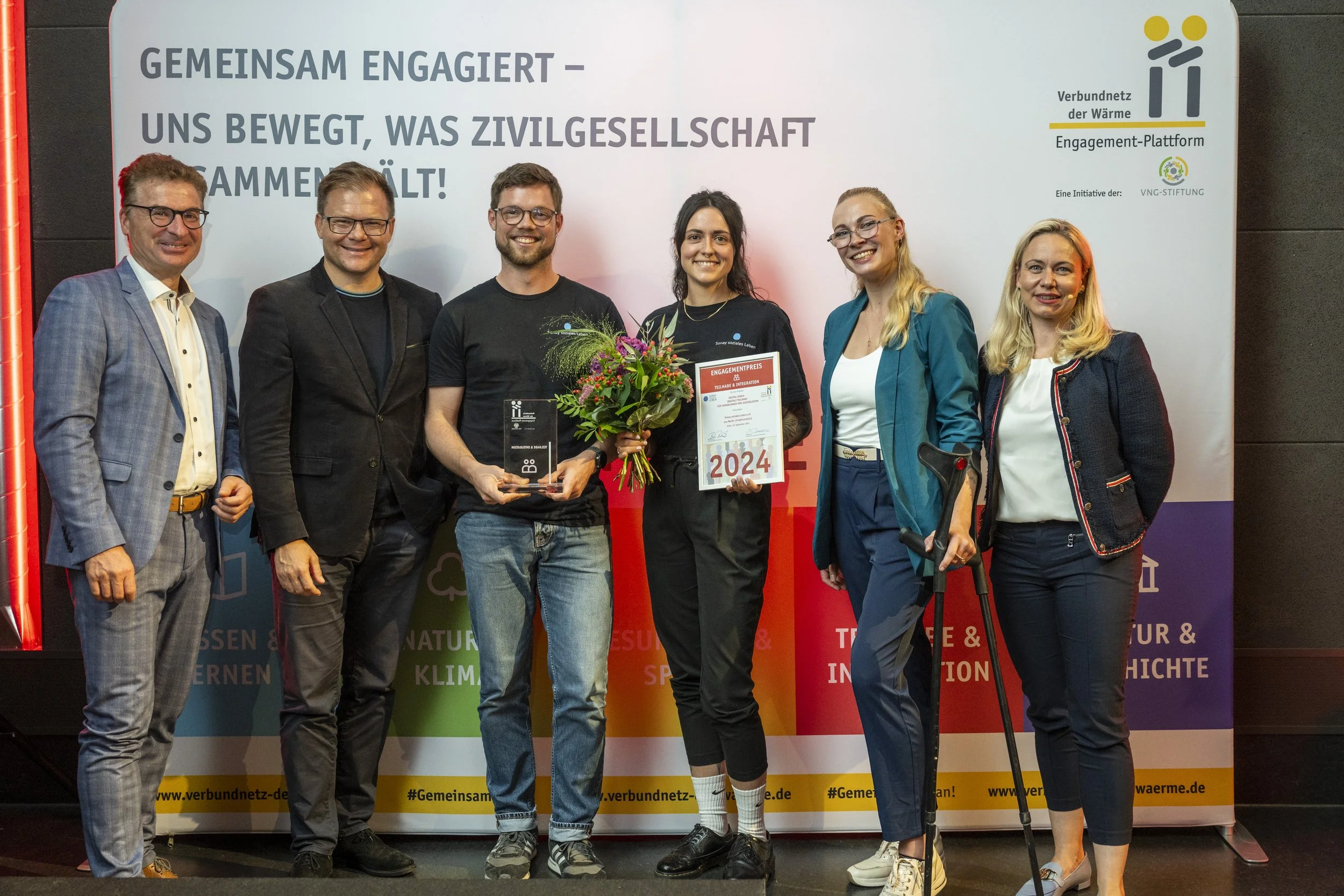 Eine Gruppe von sieben Menschen steht zusammen vor einer weißen Wand mit Text. Zwei der Frauen halten einen Blumenstrauß, eine hält eine Urkunde. Alle lächeln in die Kamera.