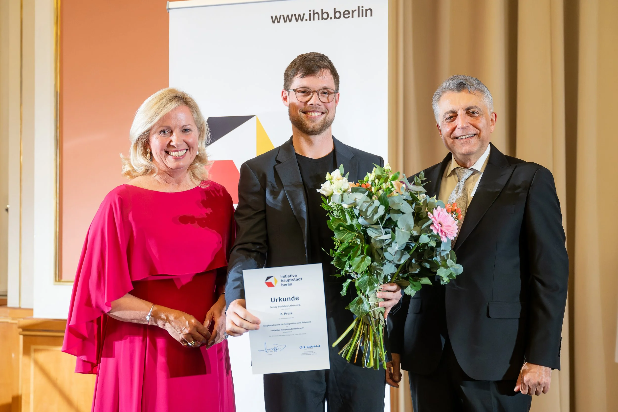 Drei Menschen bei einer Auszeichnung, im Hintergrund ein Banner mit der Webseite www.ihb.berlin, die Frau trägt ein rotes Kleid, der Mann in der Mitte hält einen Blumenstrauß und ein Zertifikat, der Mann rechts trägt einen schwarzen Anzug.
