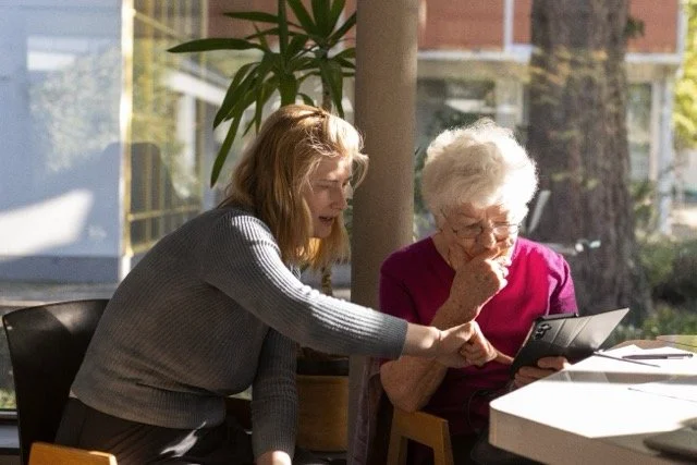 Eine junge Person zeigt einer älteren Person etwas auf dem Tablet.