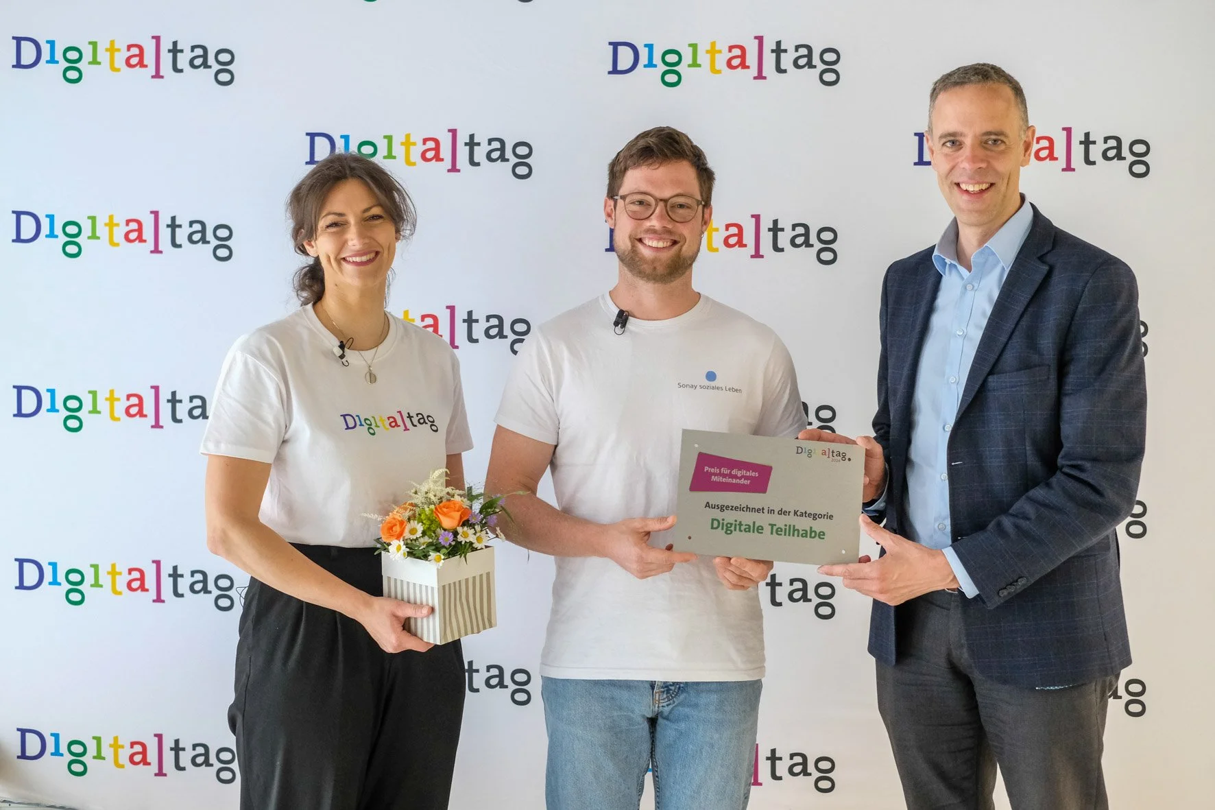 Drei Menschen bei einer Preisverleihung, die einen Preis in der Kategorie Digitale Teilhabe erhalten haben, vor einer Wand mit dem Digitaltag-Logo. Eine Frau hält einen Blumenstrauß, der Mann in der Mitte hält eine Urkunde, und der Mann auf der rechten Seite übergibt den Preis.