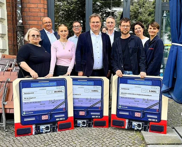 Eine Gruppe von neun Menschen steht hinter zwei große Requisiten in Form von Zügen, die als Spendenschecks gestaltet sind, auf einem gepflasterten Platz vor einem Gebäude.