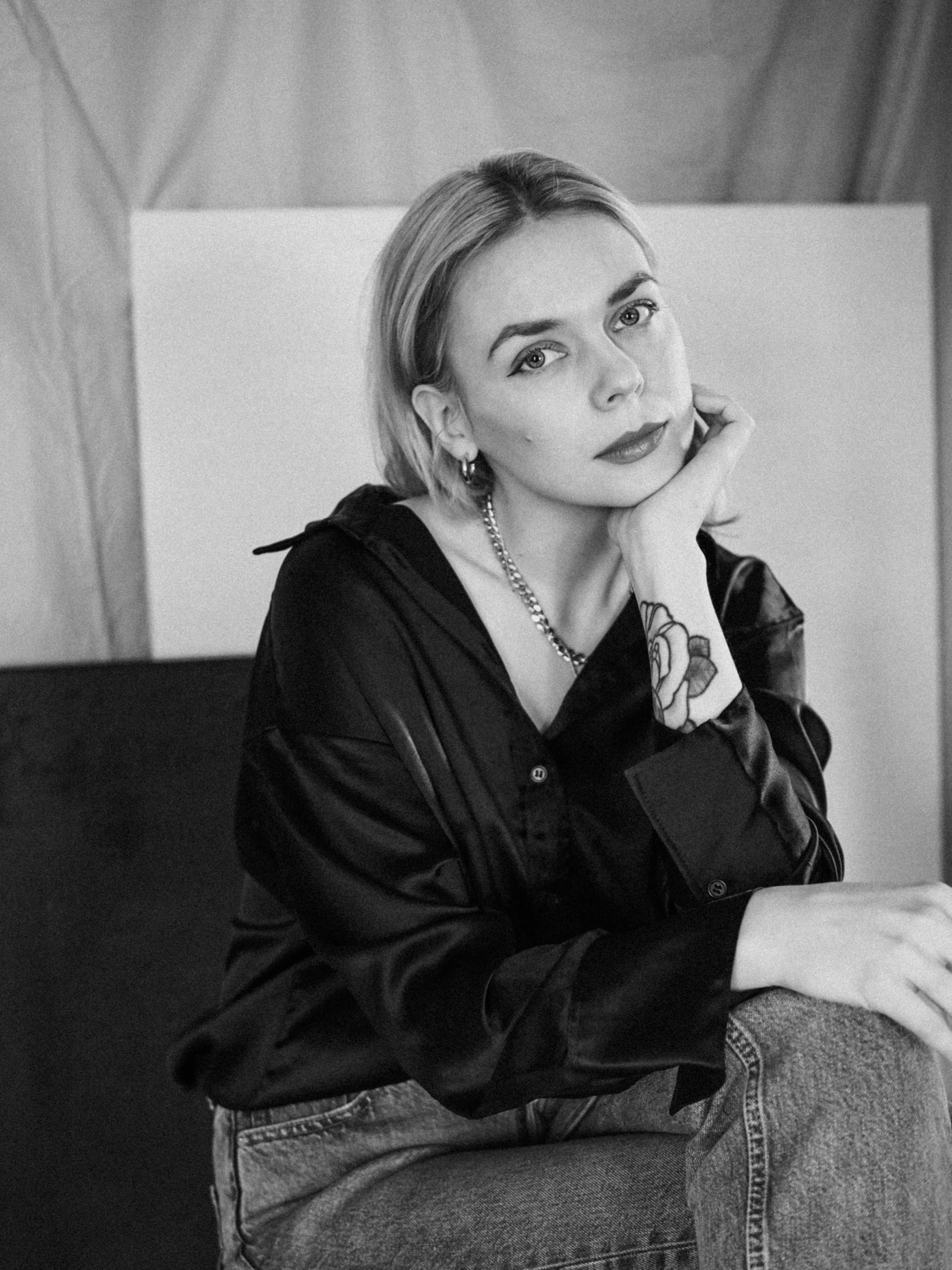 A woman with shoulder-length hair and earrings resting her face on her hand, sitting with her arm resting on her knee, dressed in a black satin jacket and jeans.