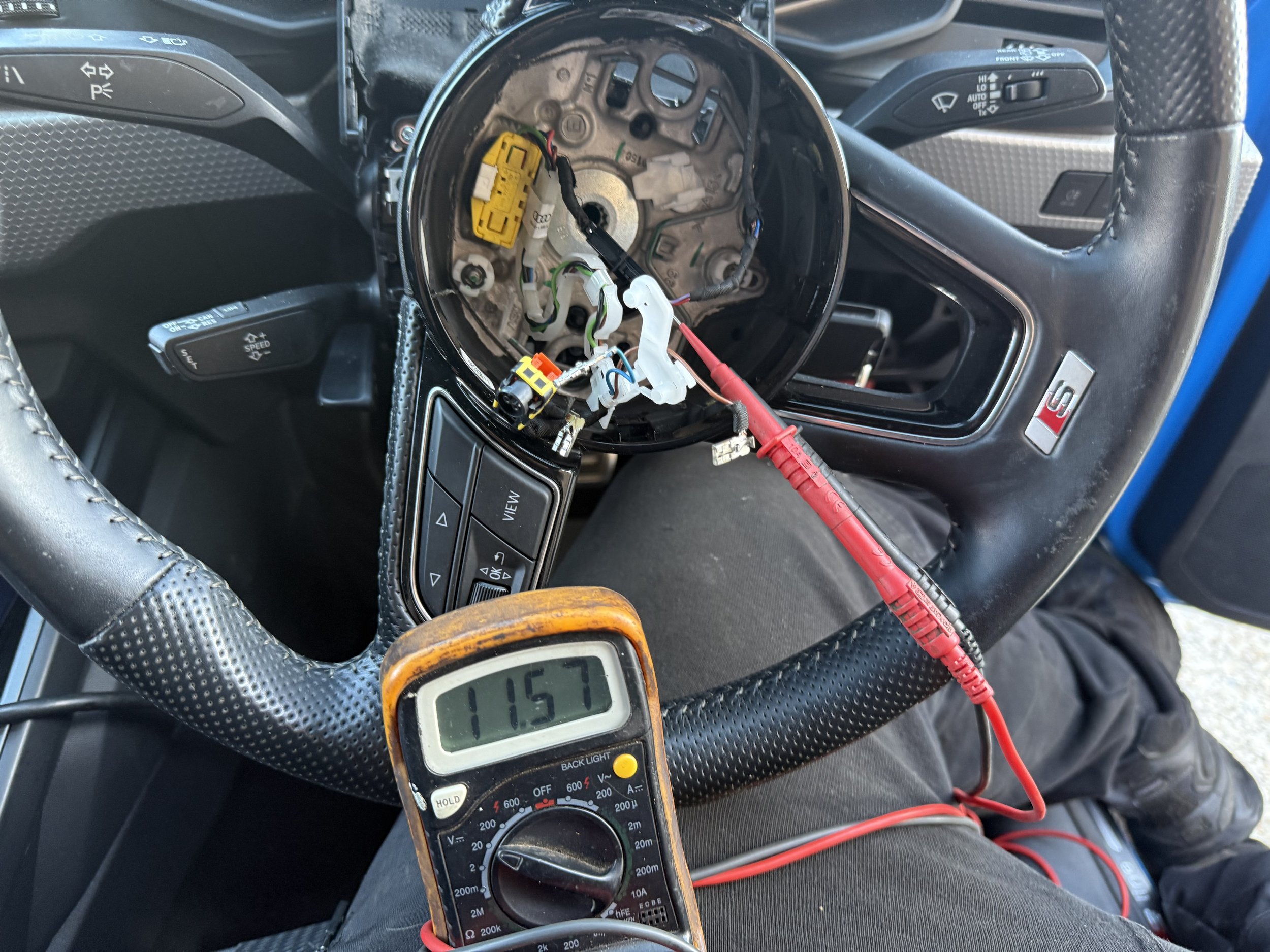 Inside a car, the dashboard and steering wheel are visible. The center console is open, revealing wiring and electronic components. A multimeter is placed on the steering wheel with a reading of 11.57. Red and black test leads are connected to the multimeter.