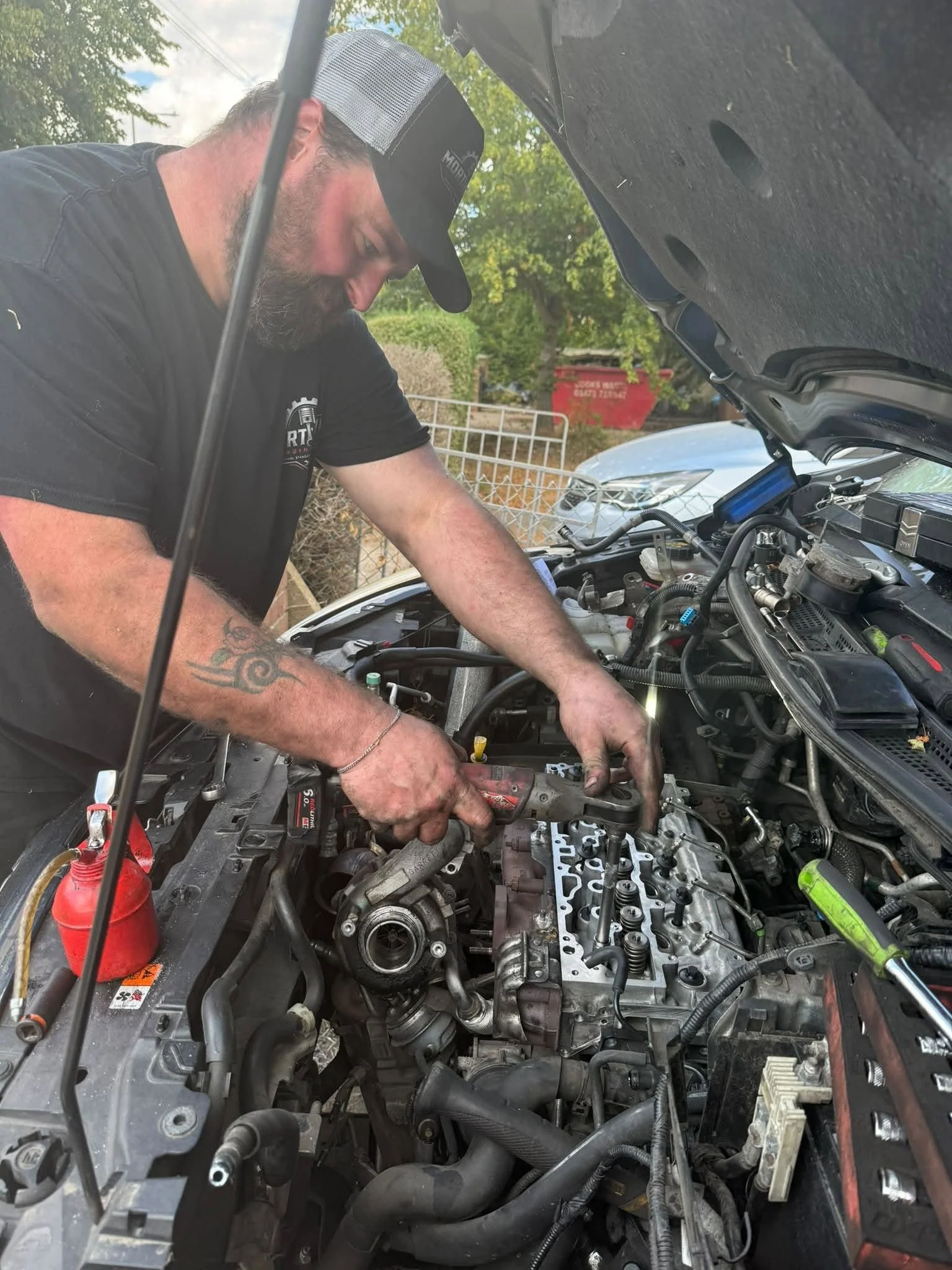 A bearded man wearing a black shirt and a black and white cap working on a car engine with the hood open, outdoors on a sunny day with trees and a fence in the background.