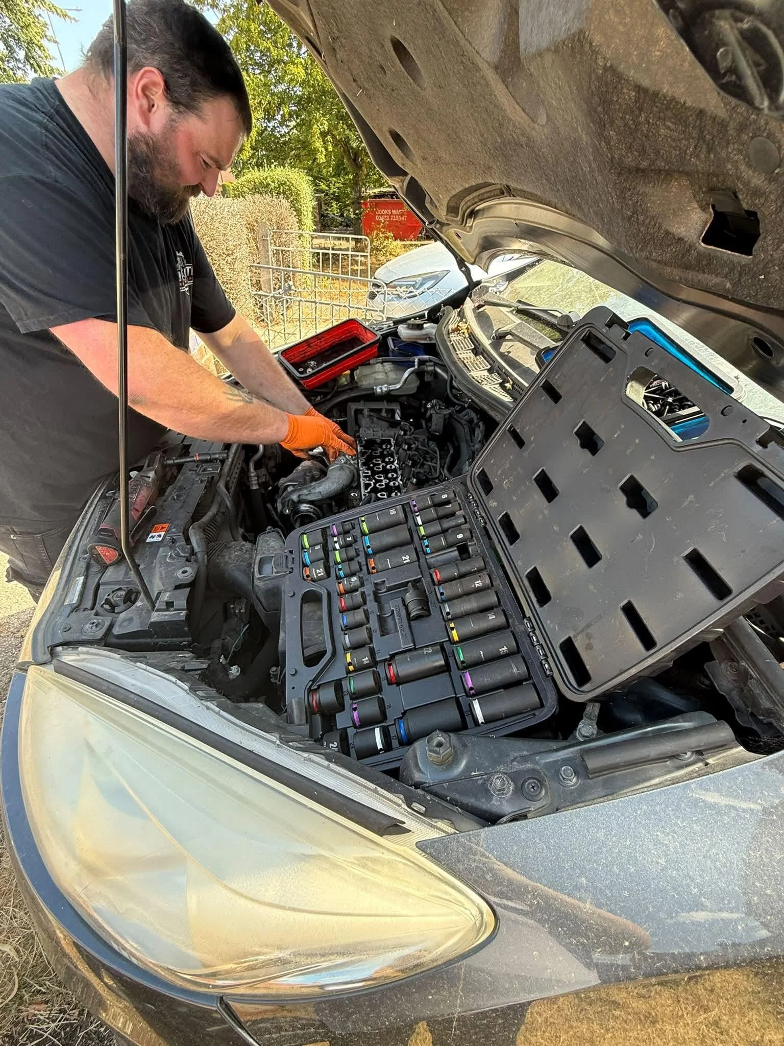 A man working on a car's engine with the hood open, surrounded by tools and a toolbox, in an outdoor setting.