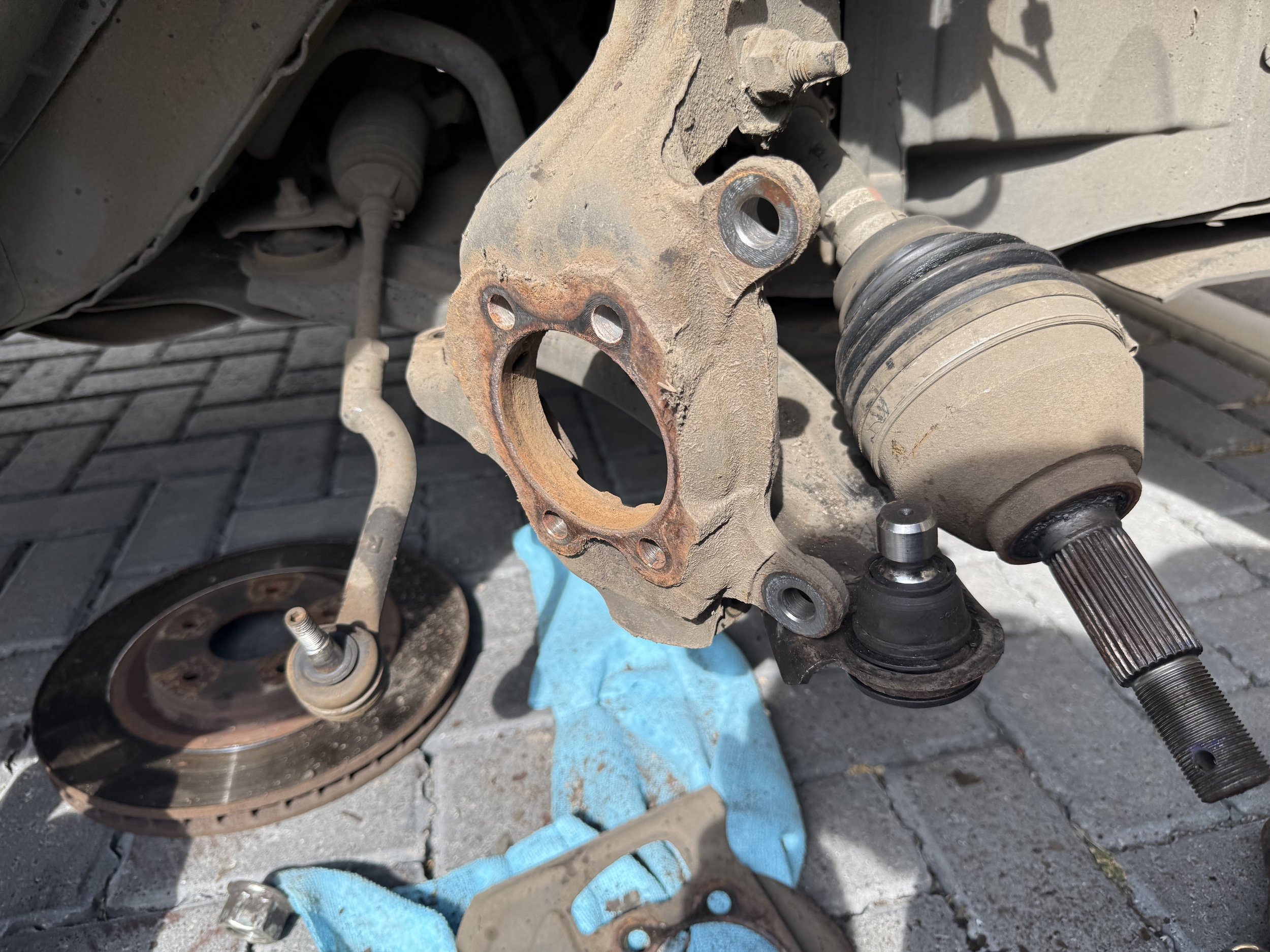 Close-up view of a car's wheel hub assembly with rusted brake rotor and missing brake caliper, exposing the wheel studs, with a blue cloth underneath and the car lifted on a jack.