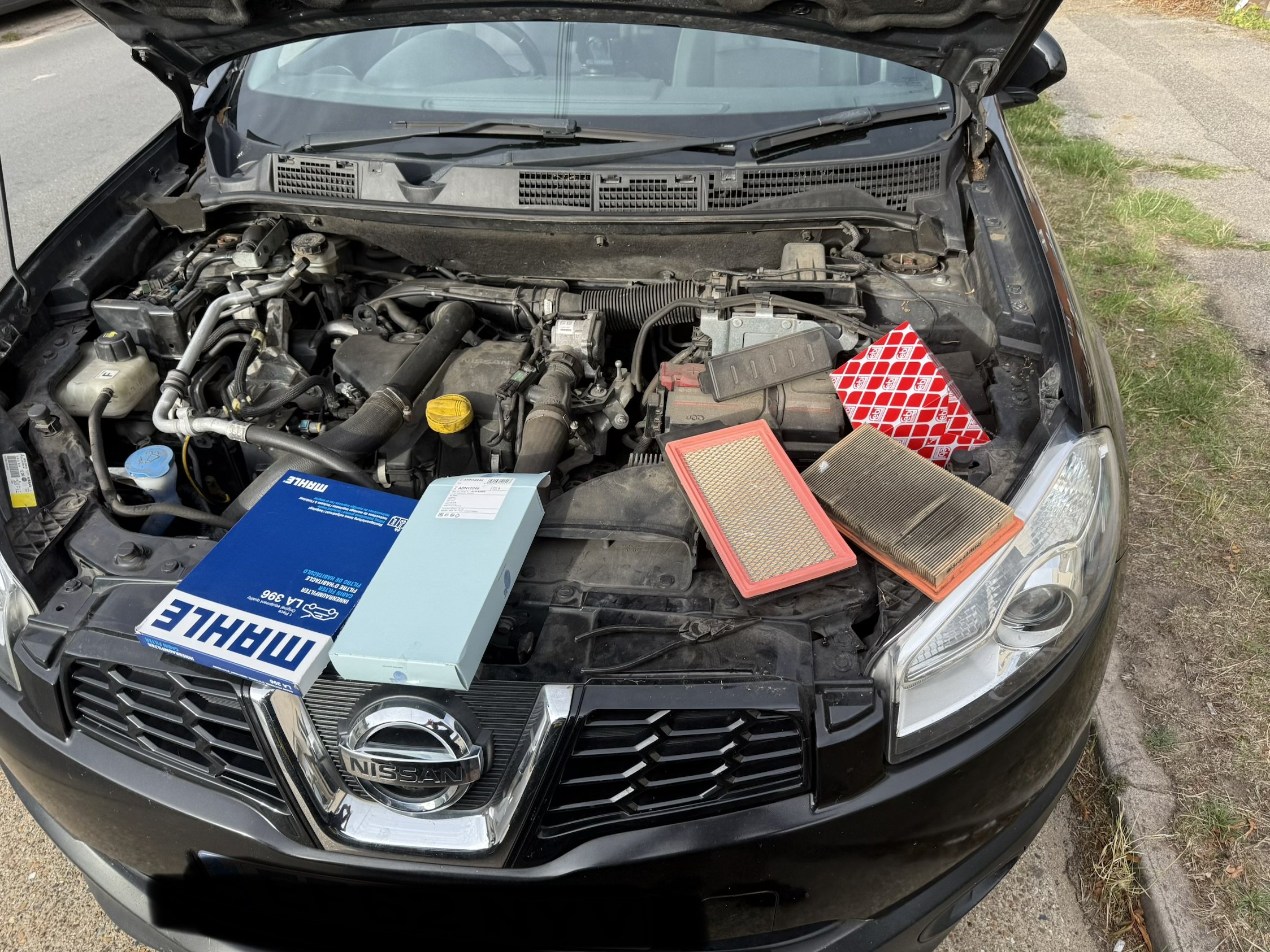 The image shows the engine bay of a black Nissan vehicle with the hood open. Various engine components and parts are visible, including a blue box labeled 'MAHLE' and a red and white air filter. The vehicle is parked on a dirt and grass surface beside a paved road.