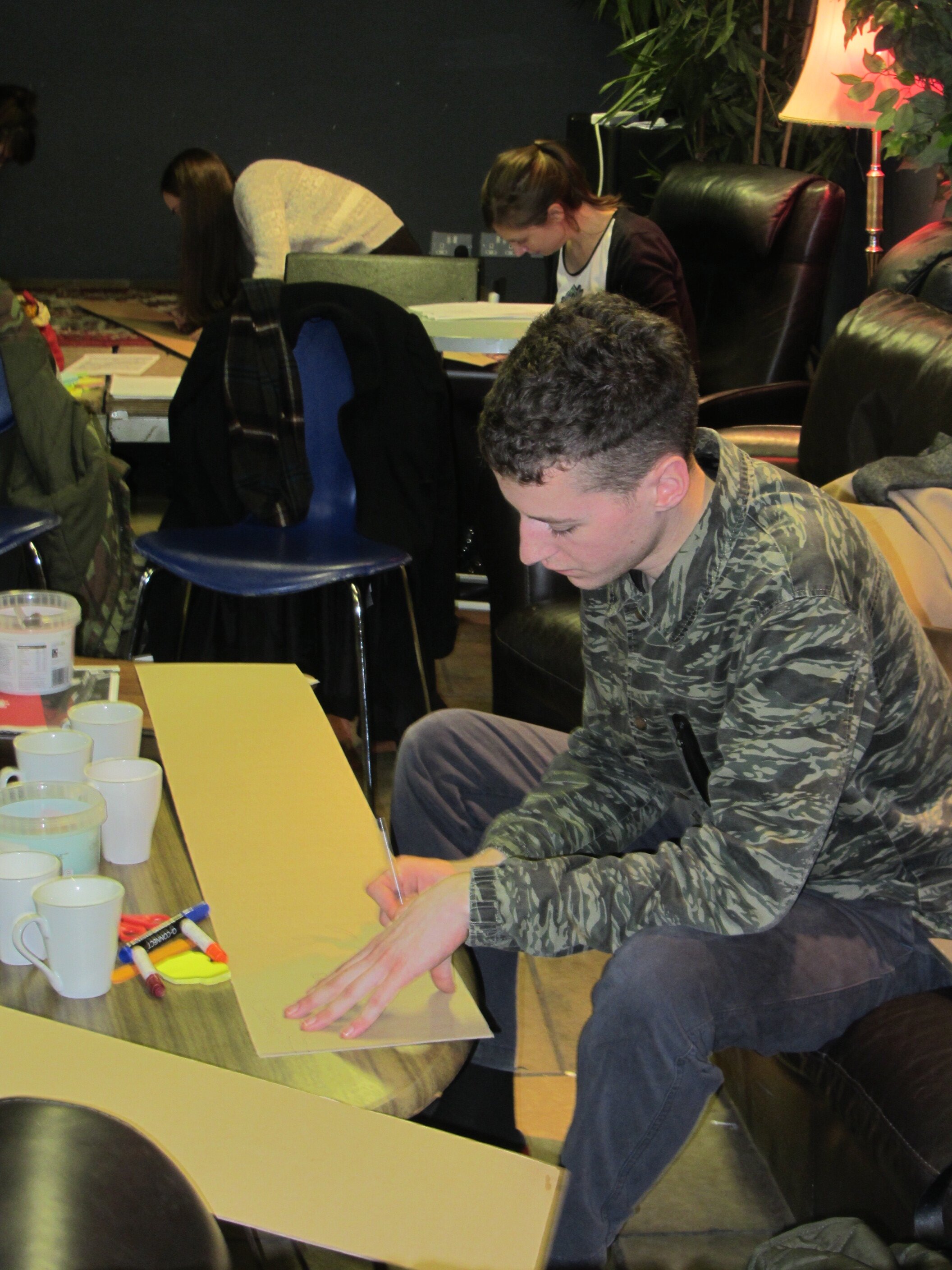 A man is sitting on a couch, cutting a piece of cardboard.