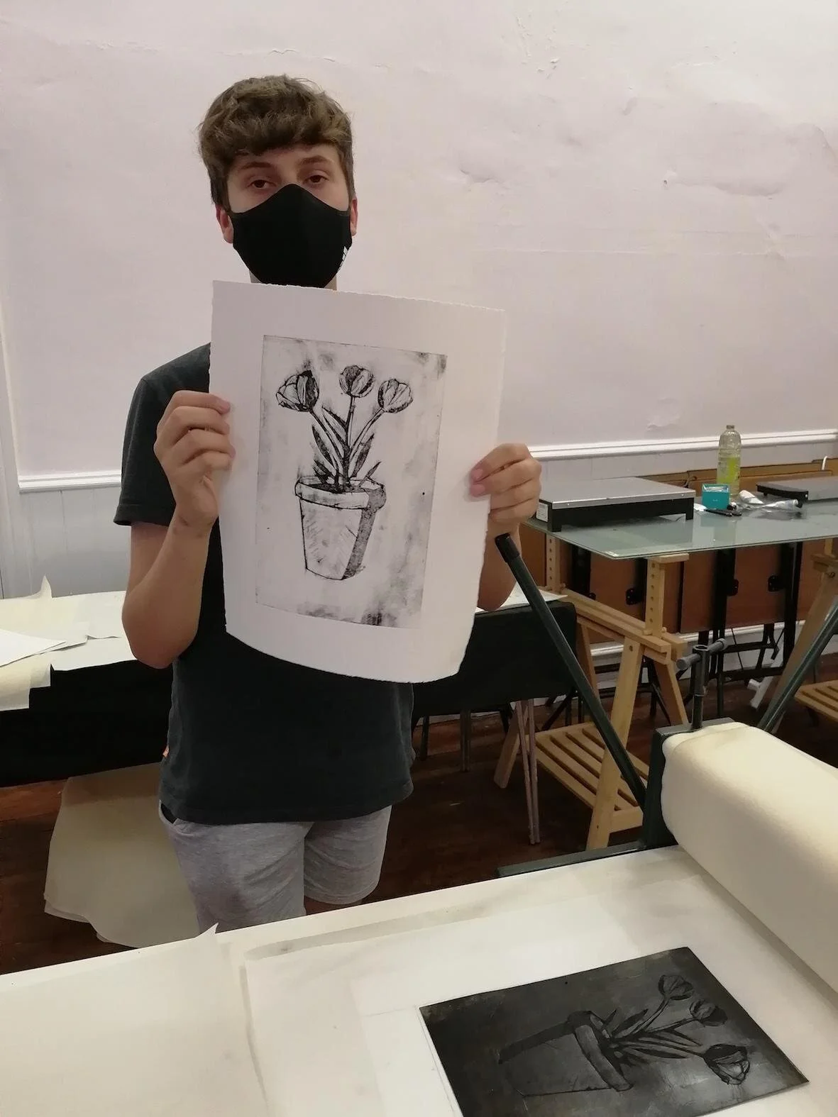 A young person in a black face mask holds up a black and white print of tulips in a pot, in a printmaking studio at a Create Glos workshop.