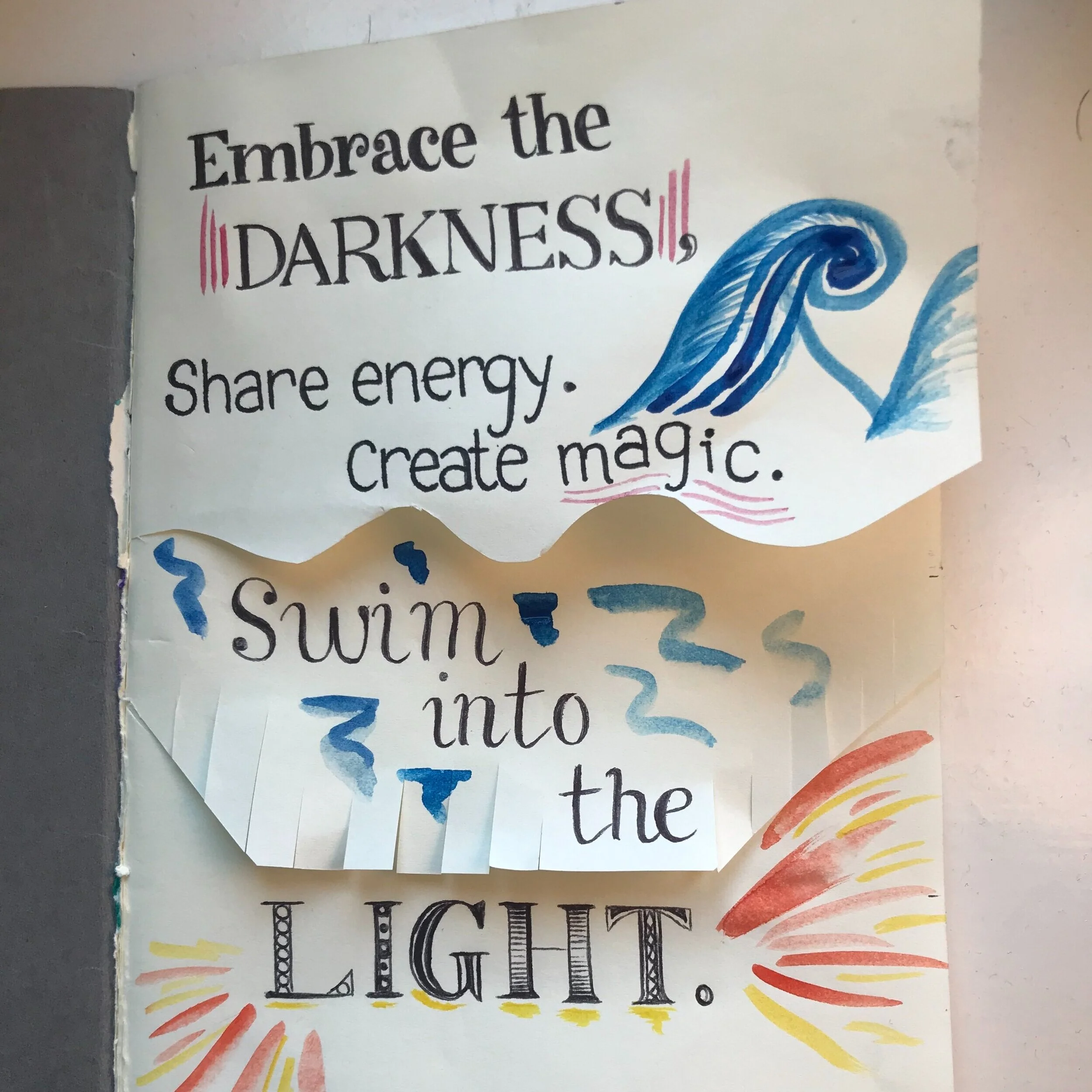 Handwritten artwork reading “Embrace the darkness, share energy, create magic. Swim into the light”, with blue waves.