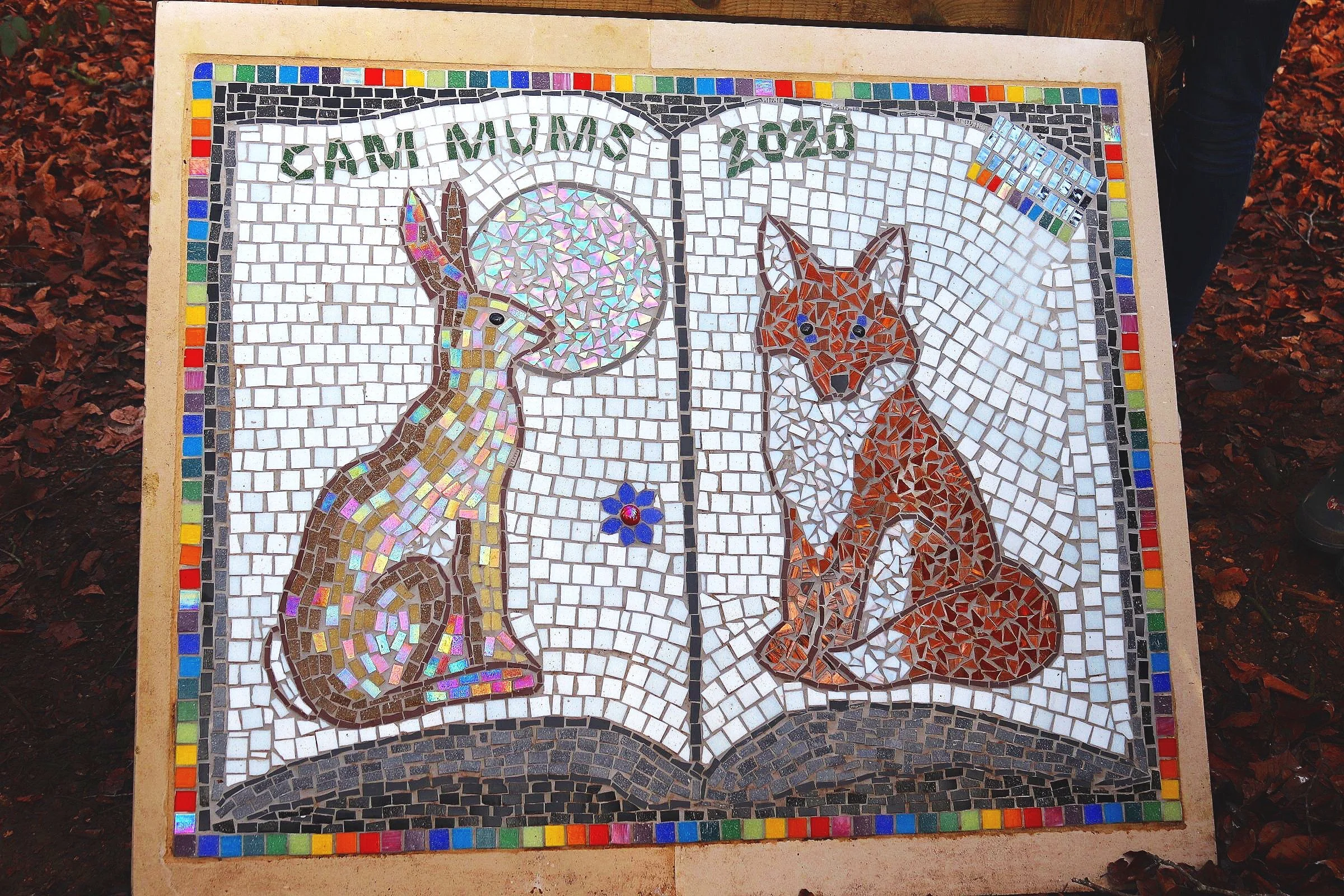 A photo of a sculpture which has been designed to look like an open book. On the left hand side there is a rabbit and on the right a fox. It is made using mosaic tiles and there is a title at the top that reads' cam mums 2020'