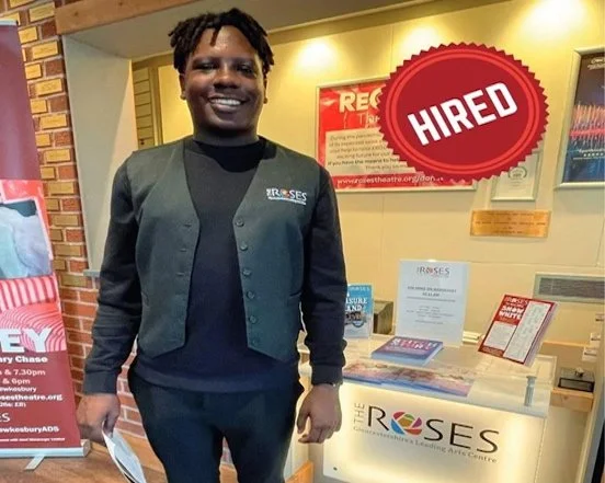 Jordan is wearing a Roses Theatre waistcoat and smiling at a reception desk in the foyer of the theatre.