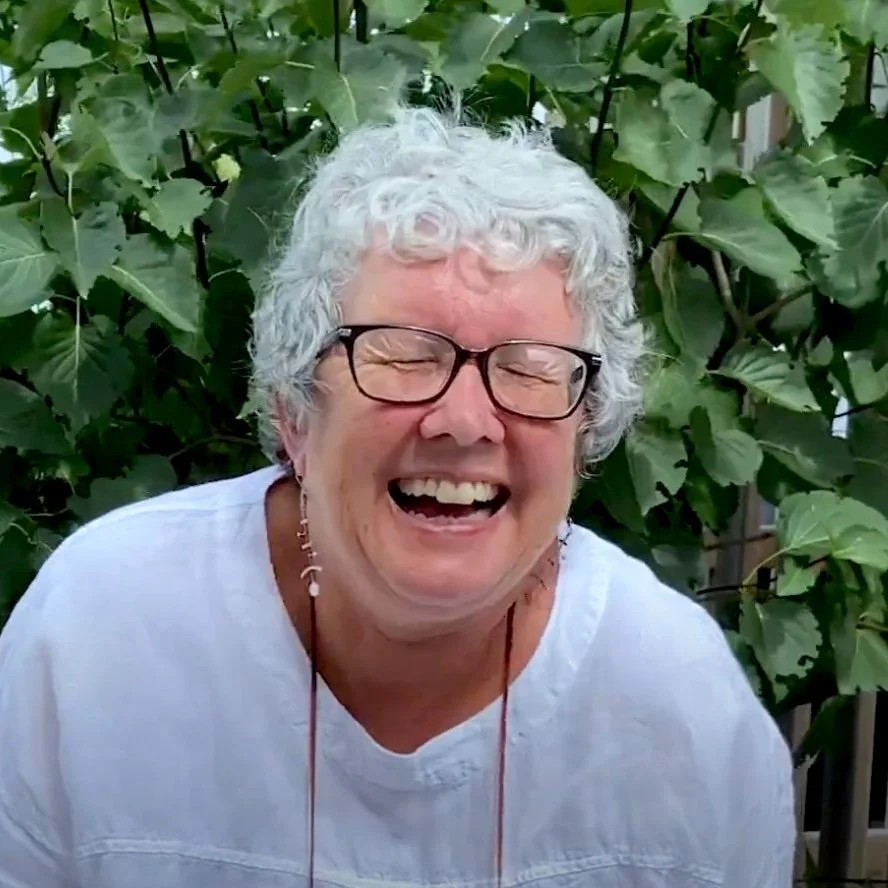 Karen, laughing, with short grey hair and glasses, wearing a white top, with green leaves behind her.