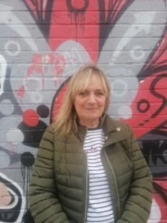 Debbie, with blonde hair, wearing a green jacket and striped top, standing in front of a red and grey mural.