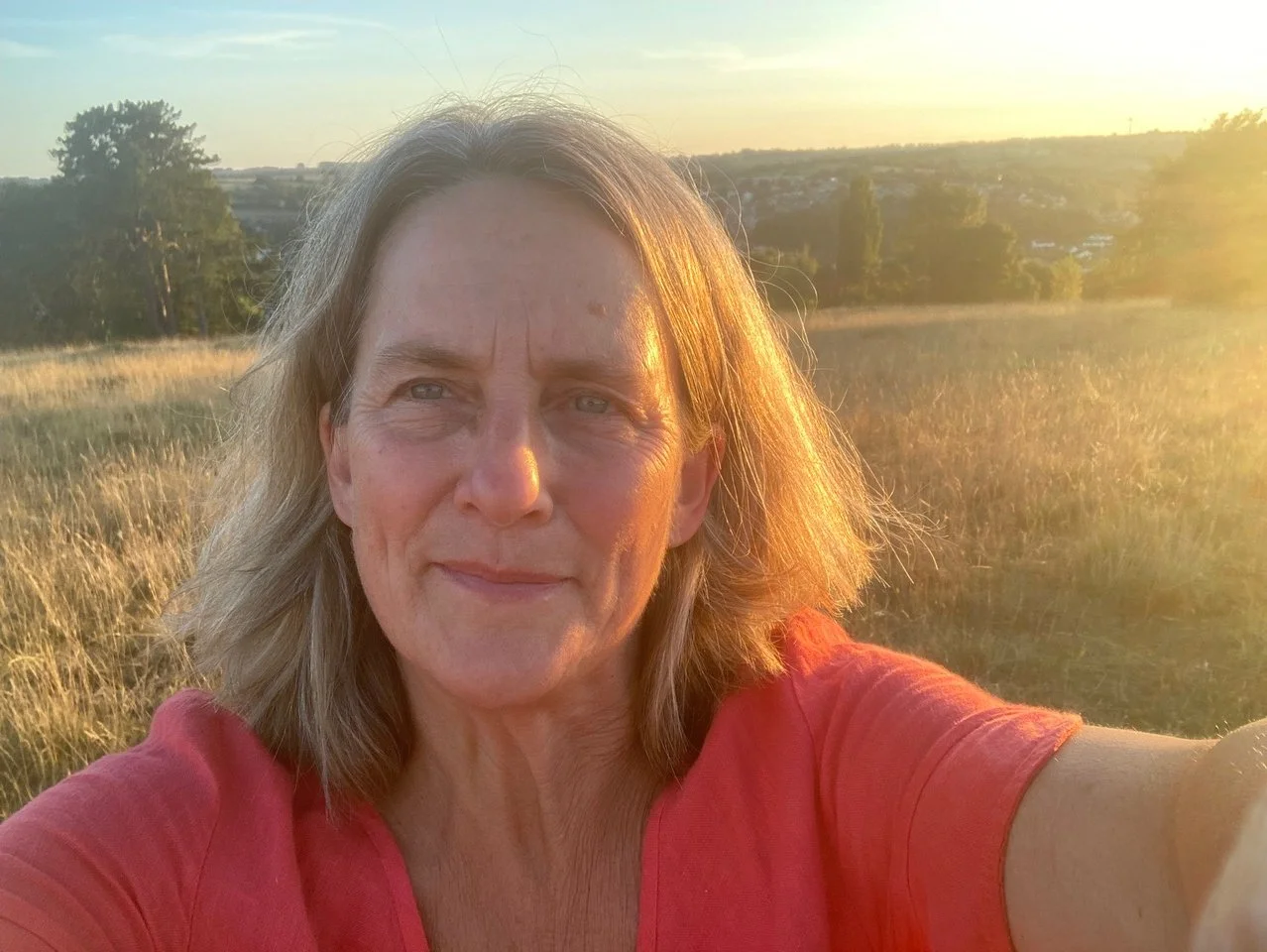 Pippa, smiling in a red top, taking a selfie in a golden field at sunset.