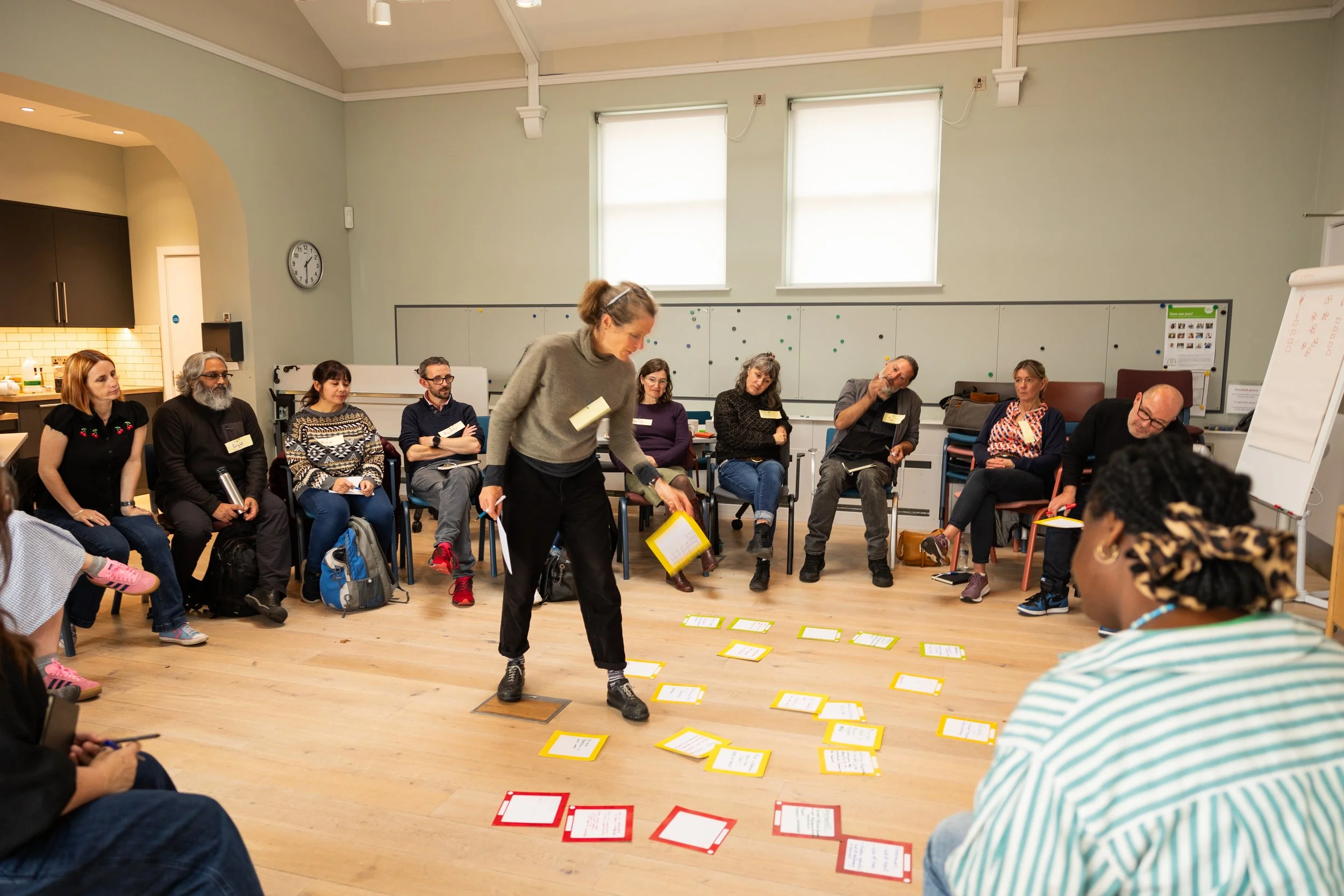 A woman stands in the middle of a circle of seated people, laying out red-bordered cards on the floor during a Create Glos event.