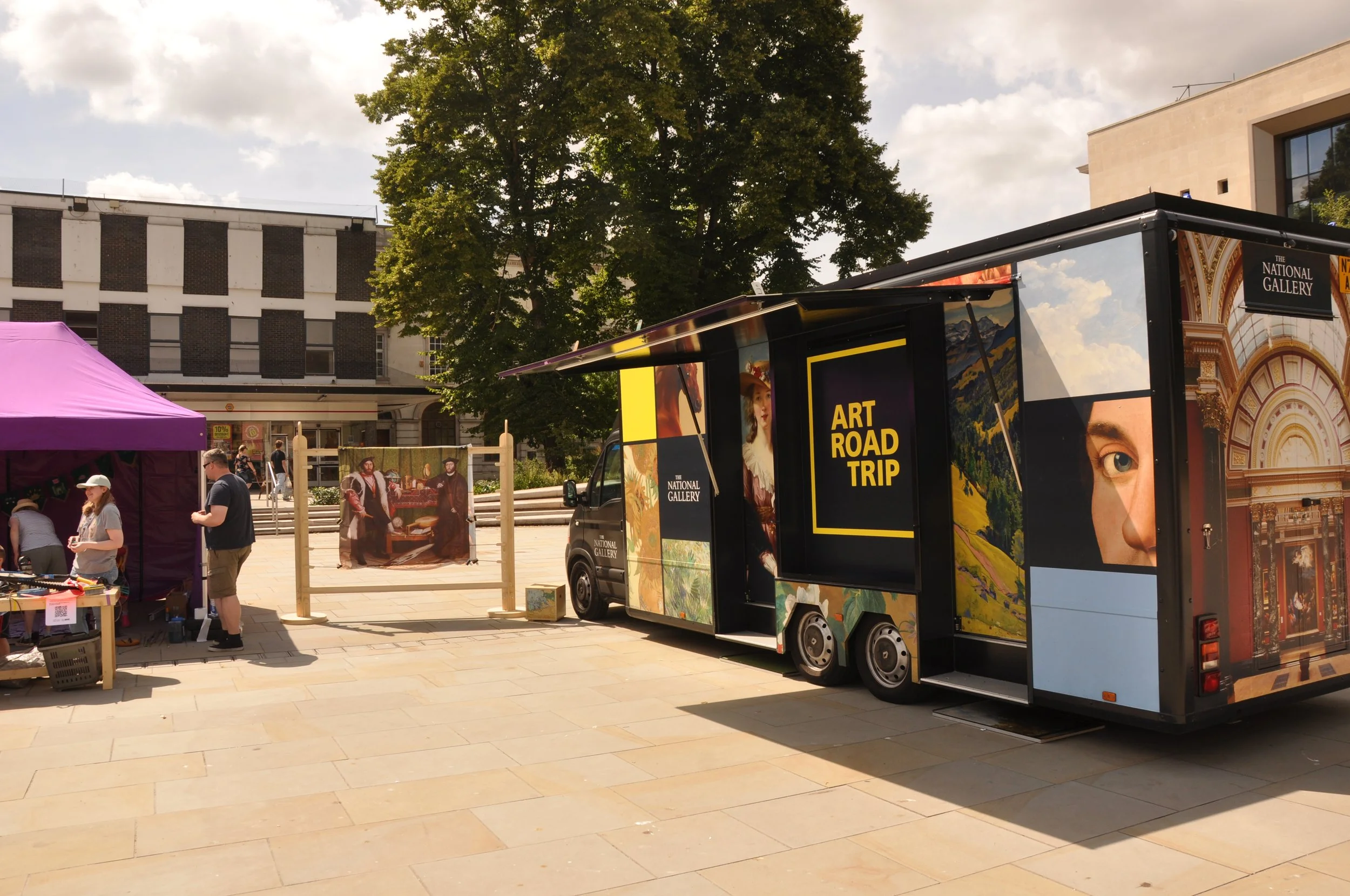 The National Gallery Art Road Trip van in a public square with Holbein's The Ambassadors painting displayed next to a purple gazebo