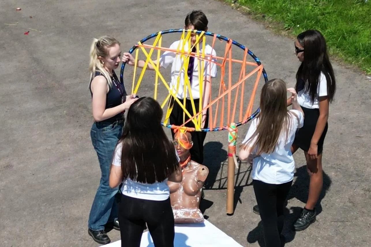 Four school children and one arts facilitator hold a circular sculpture with orange and yellow ribbon in a playground space