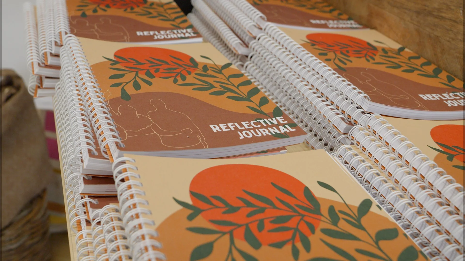 Stacks of spiral-bound Reflective Journals with orange sun and green leaf artwork by Lucy Turner.