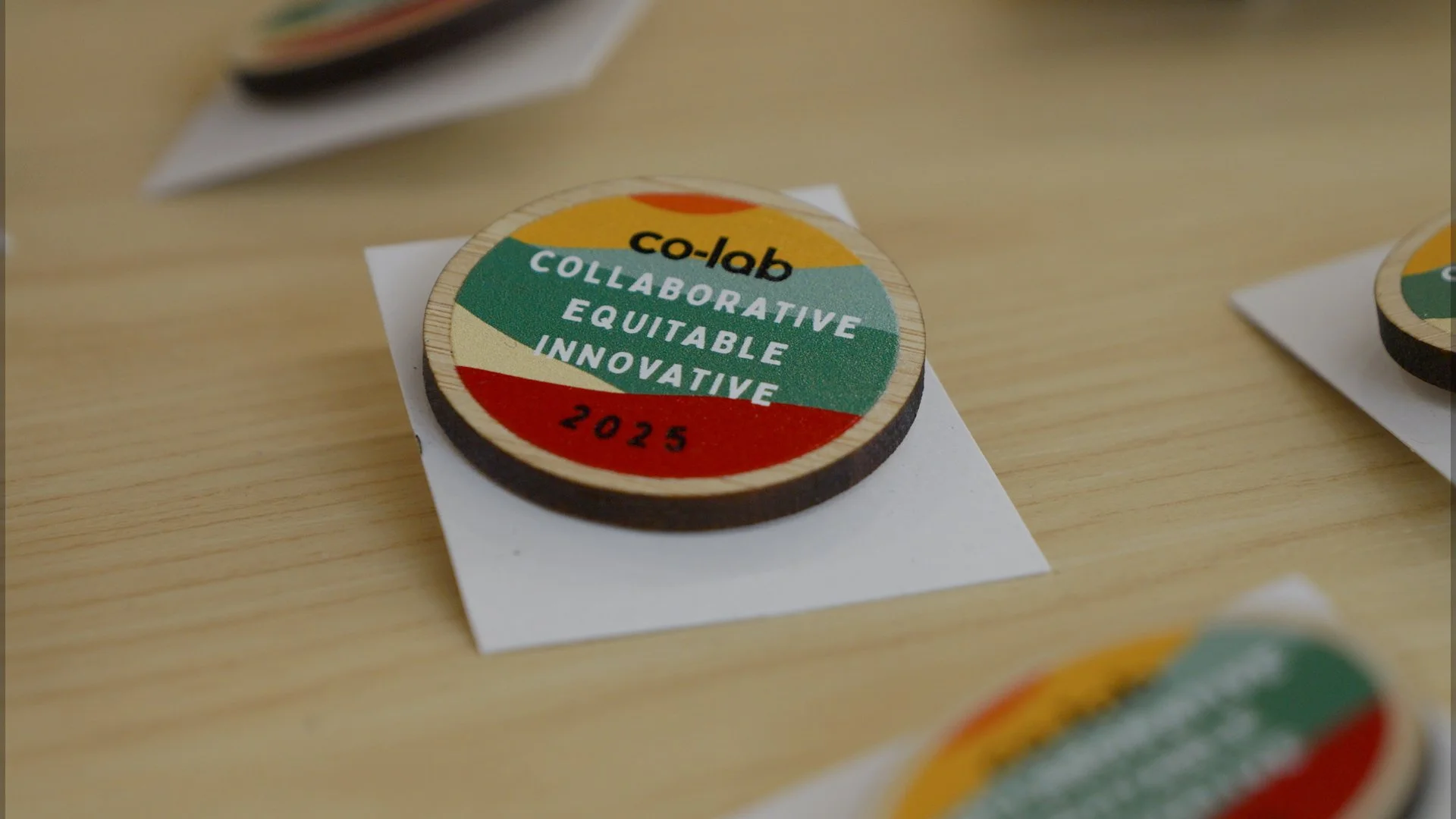 Round wooden co-lab 2025 badge reading “Collaborative, Equitable, Innovative”, with colourful artwork by Lucy Turner.