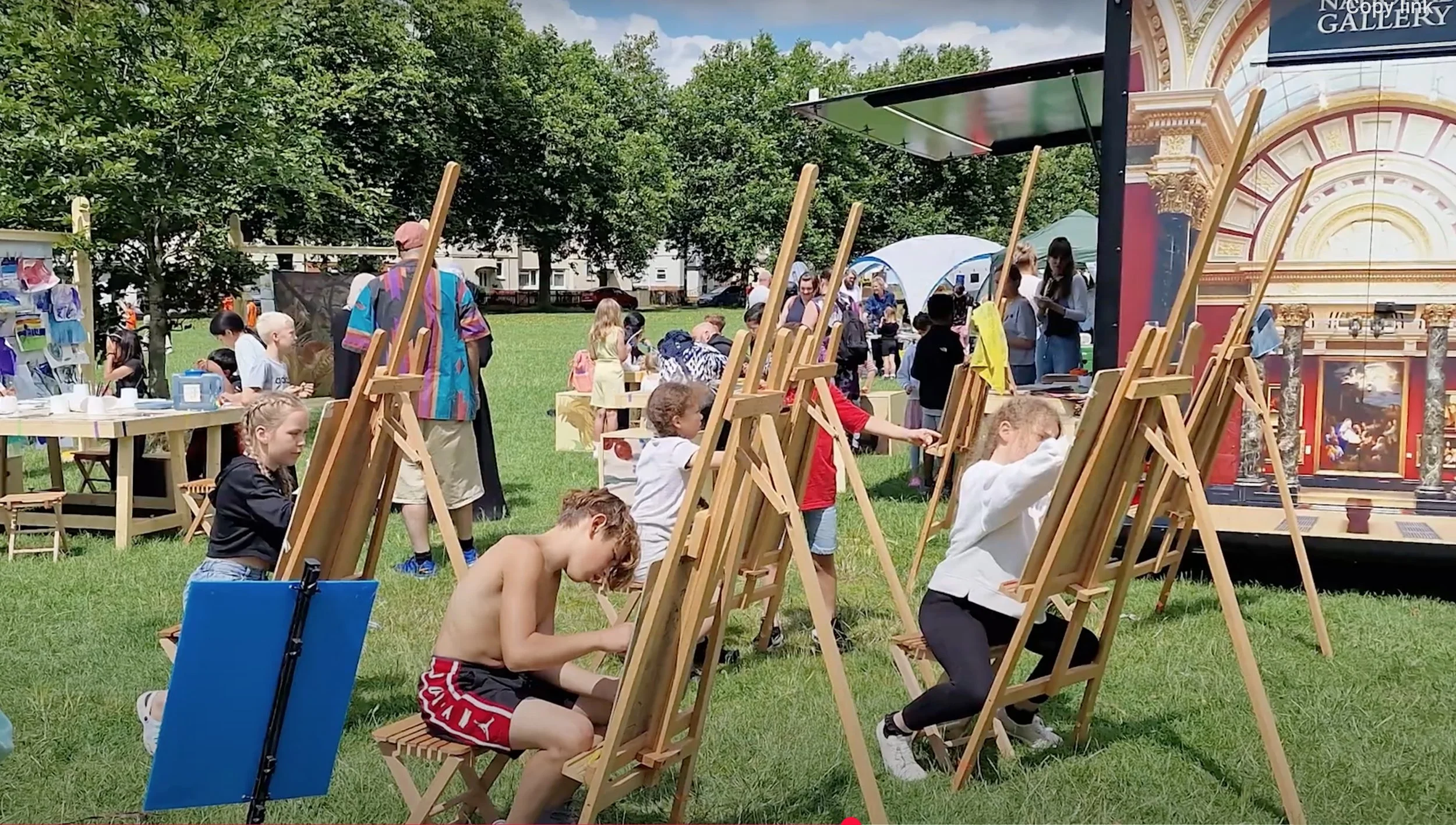 People paint at wooden easels on a sunny lawn, at an outdoor Create Glos art event.