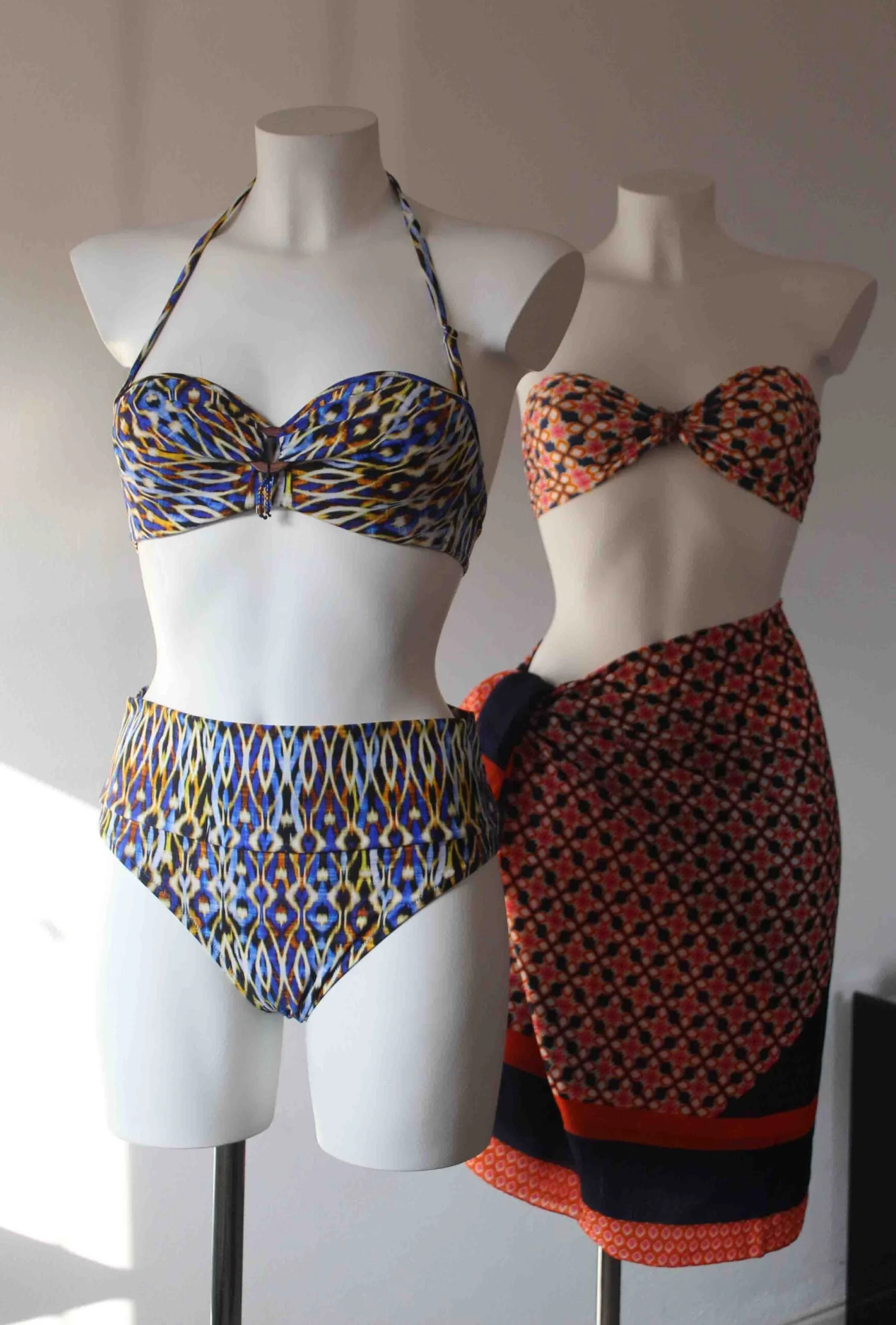 Two white mannequins display patterned bikinis; the left mannequin has a blue and yellow abstract design, the right mannequin has a floral pattern with black, orange, and pink colors.