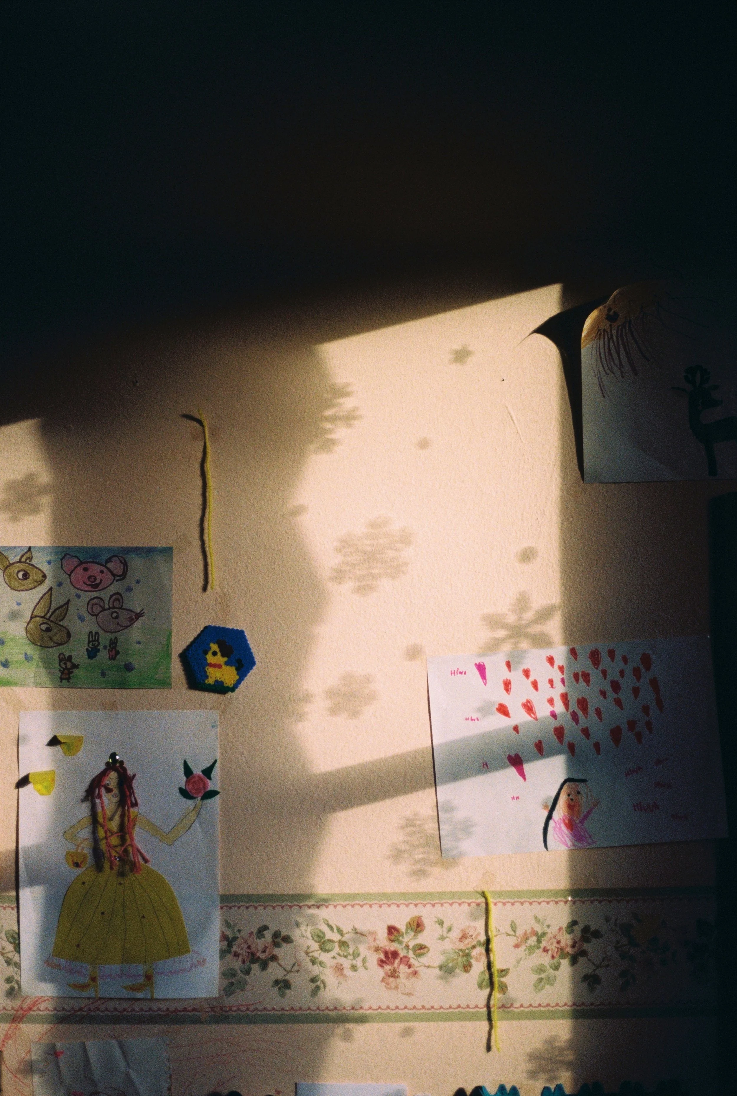 Zeichnungen und Collagen von Kindern auf einer Wand, darunter ein Papier mit einem Märchenprinzen und eine Karte mit Herzen, Schatten von Pflanzen an der Wand.