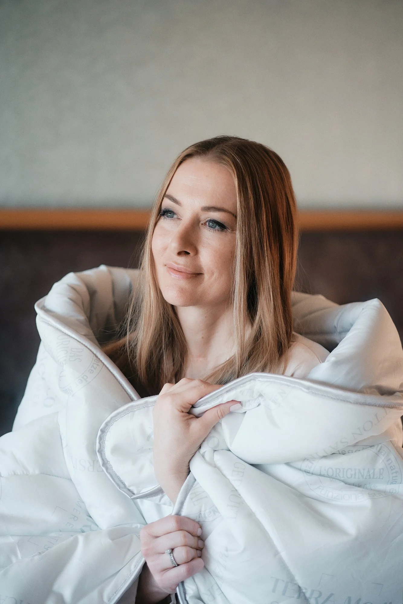 Eine Frau mit langen braunen Haaren sitzt auf einem Sofa, trägt ein weißes Oberteil und lächelt sanft. Sie hält eine weiße Decke um ihre Schultern, die mit Mustern und Text bedruckt ist.