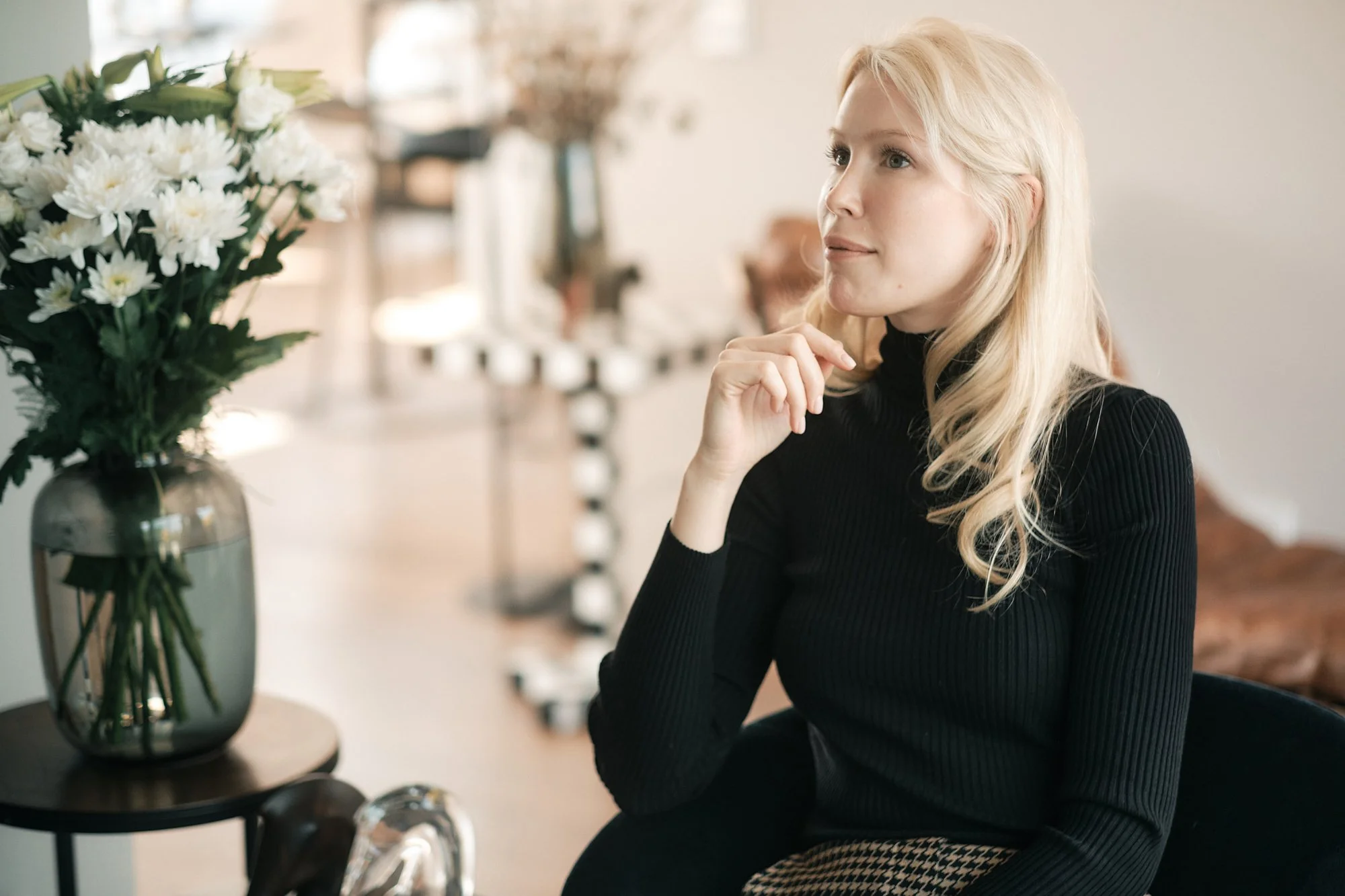 Junge blonde Frau sitzt im Raum neben einem Tisch mit einer großen Vase voller weißer Blumen, trägt ein schwarzes Oberteil, scheint nachdenklich oder interessiert zu sein.