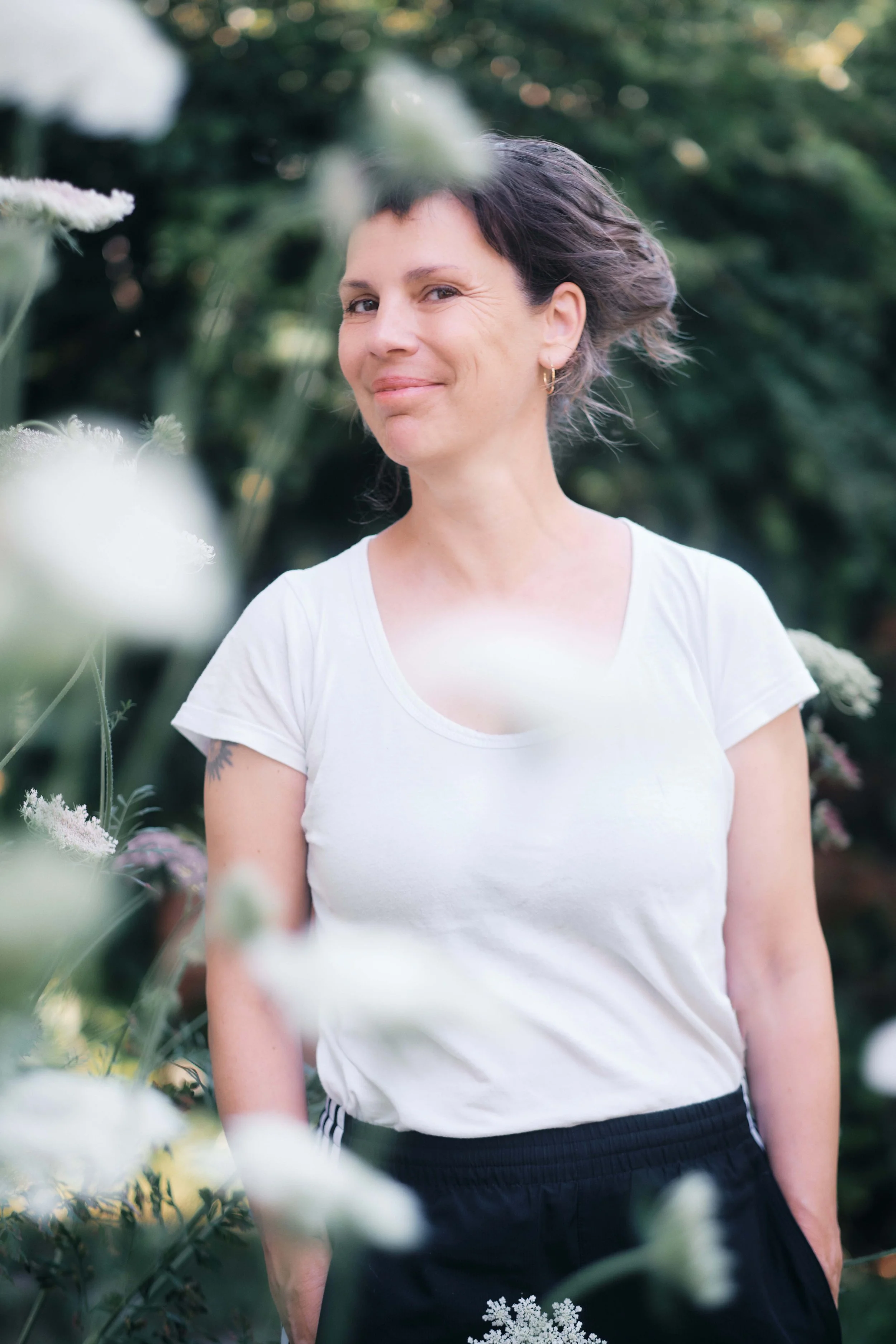 Eine lachende Frau mit kurzen dunklen Haaren, tragt ein weißes T-Shirt und eine schwarze Hose, steht im Grünen, umgeben von Blumen und Pflanzen.