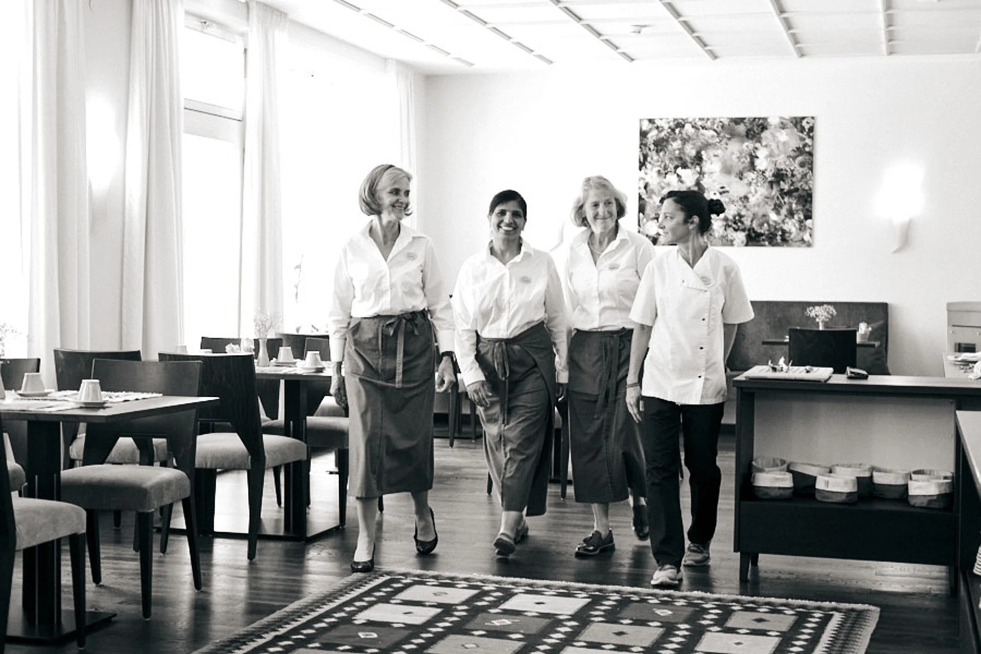 Vier Frauen in weißen Oberteilen und schwarzen Schürzen gehen durch einen hell erleuchteten Raum in einem Restaurant. Der Raum ist mit Tischen, Stühlen und einer Wand mit einem Blumenbild im Hintergrund ausgestattet.