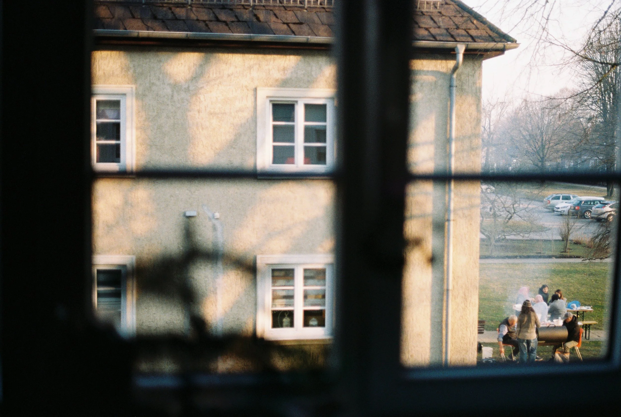 Blick durch ein Fenster auf ein Wohnhaus und einen Garten mit Menschen, die dort spazieren gehen oder zusammensitzen, bei Sonnenuntergang.