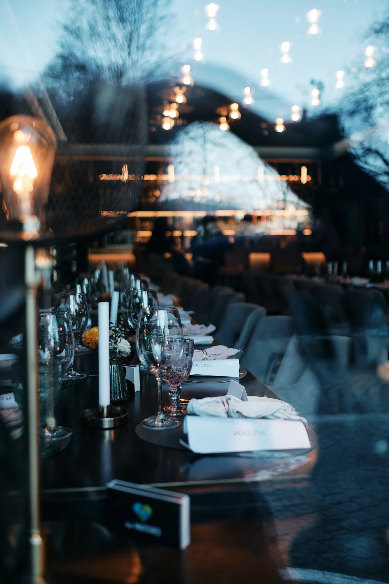 Abendessen-Tisch mit Weingläsern, Kerzen, Blumen und Besteck in einem eleganten Restaurant mit hoher Decke und großen Fenstern.