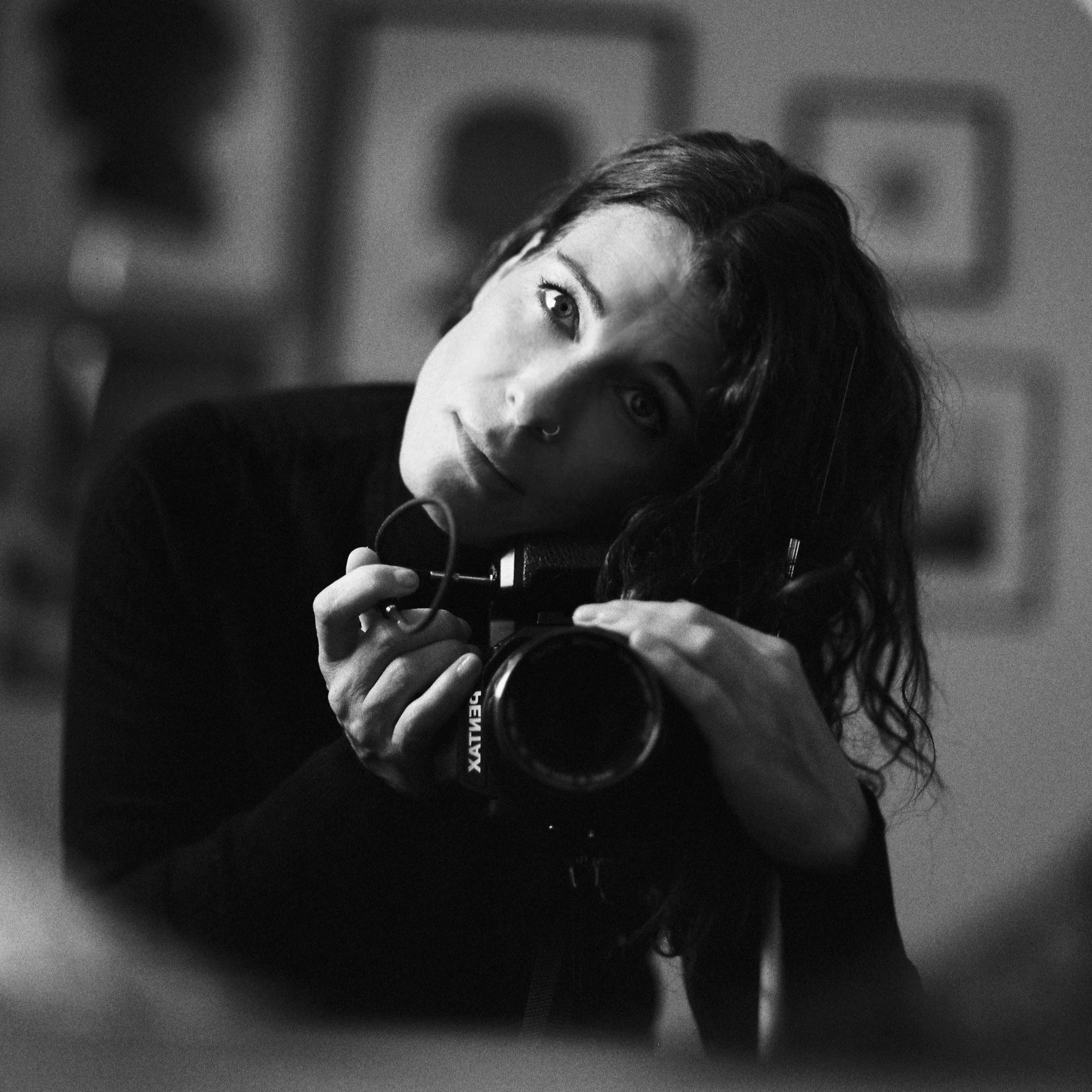 Schwarz-weiß-Fotografie einer jungen Frau mit dunklen, lockigen Haaren, die eine Kamera hält und nach oben blickt.