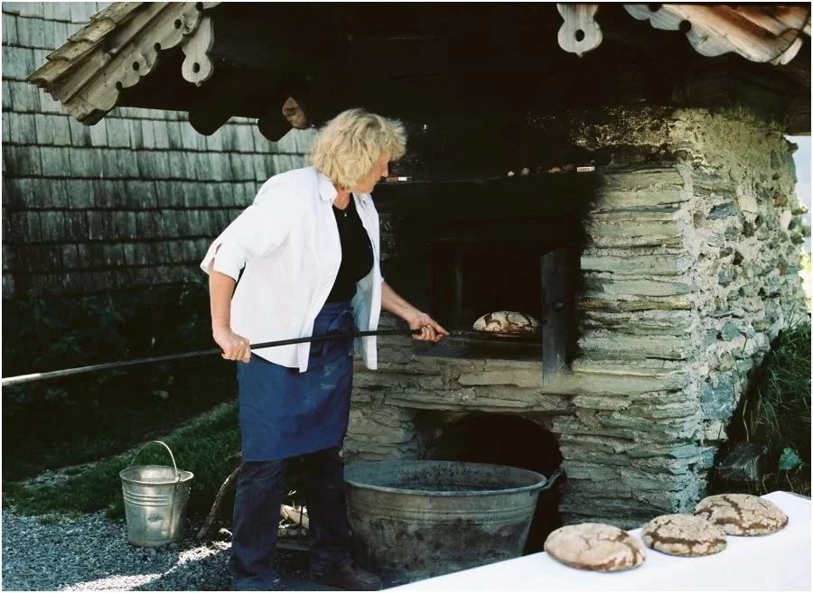 Eine Frau backt in einem Steinofen, der im Freien steht, mit mehreren frisch gebackenen Brötchen vor einem Tisch