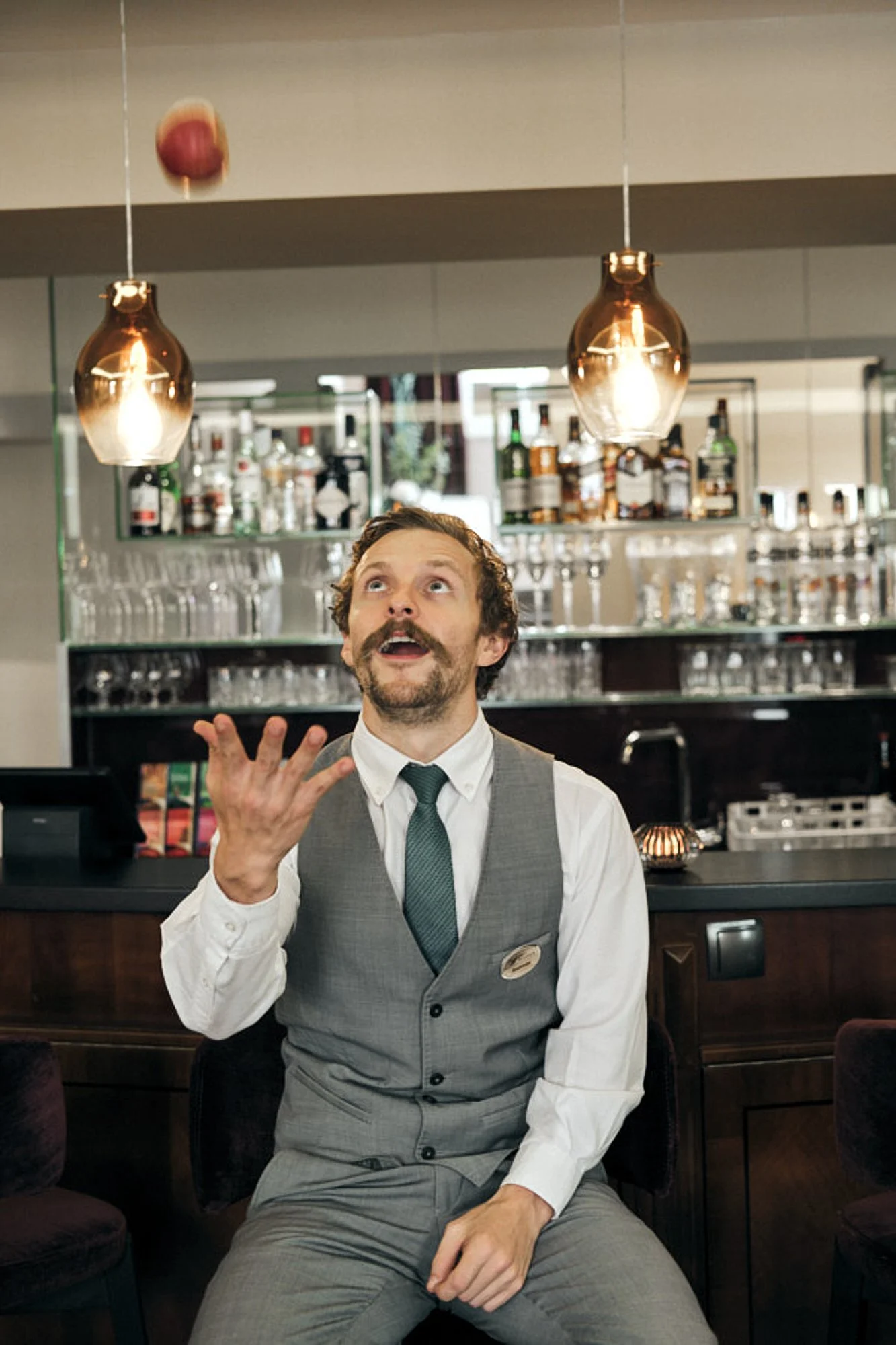Ein Mann im Anzug sitzt in einer Bar und spricht, während er seine Hand hebt. Im Hintergrund sind Regale mit Flaschen und Gläsern, die von warmem Licht beleuchtet werden.