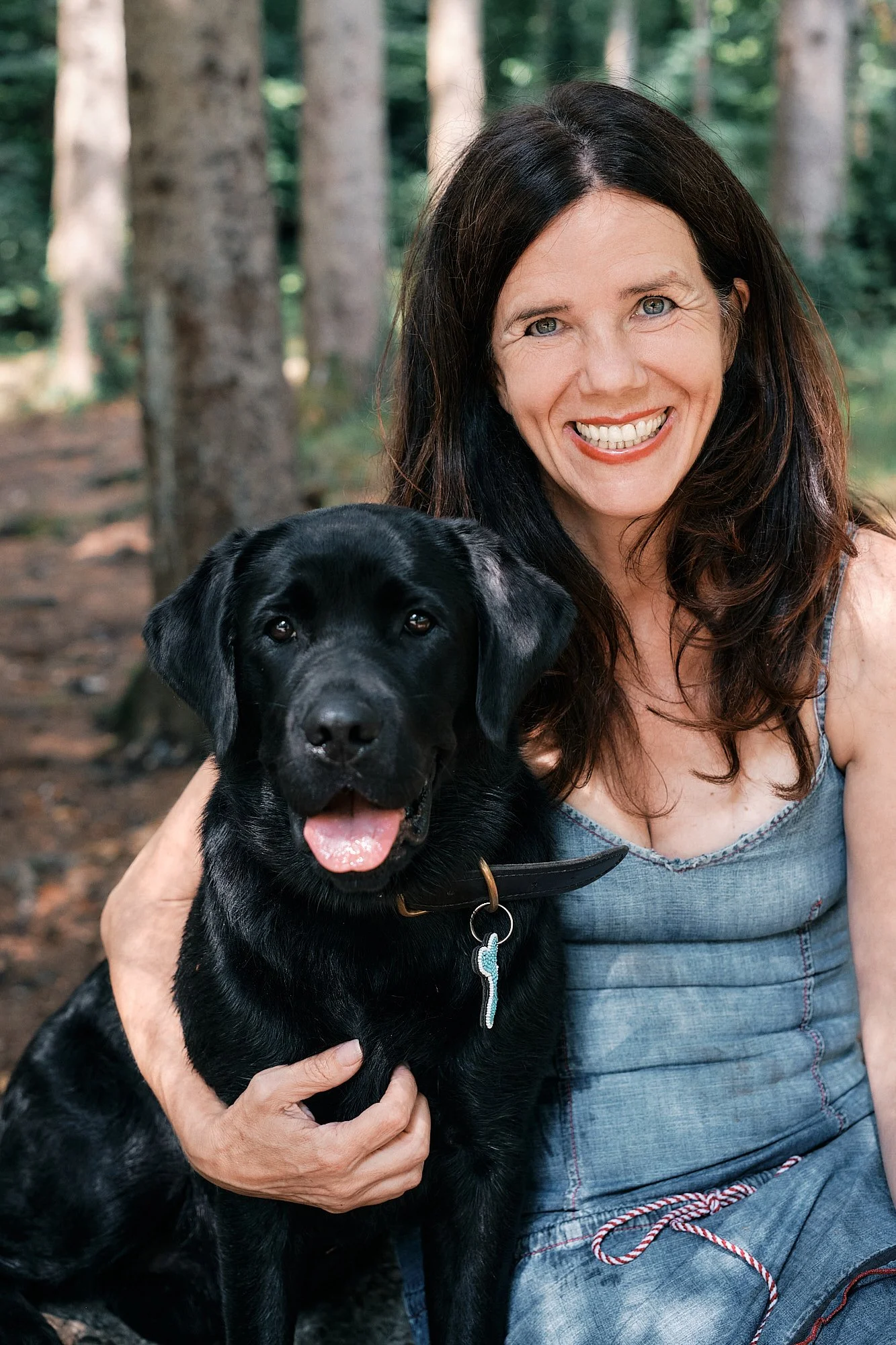 Frau mit schwarzen Haaren und Lächeln, hält schwarzen Hund mit offener Zunge in einem Wald.