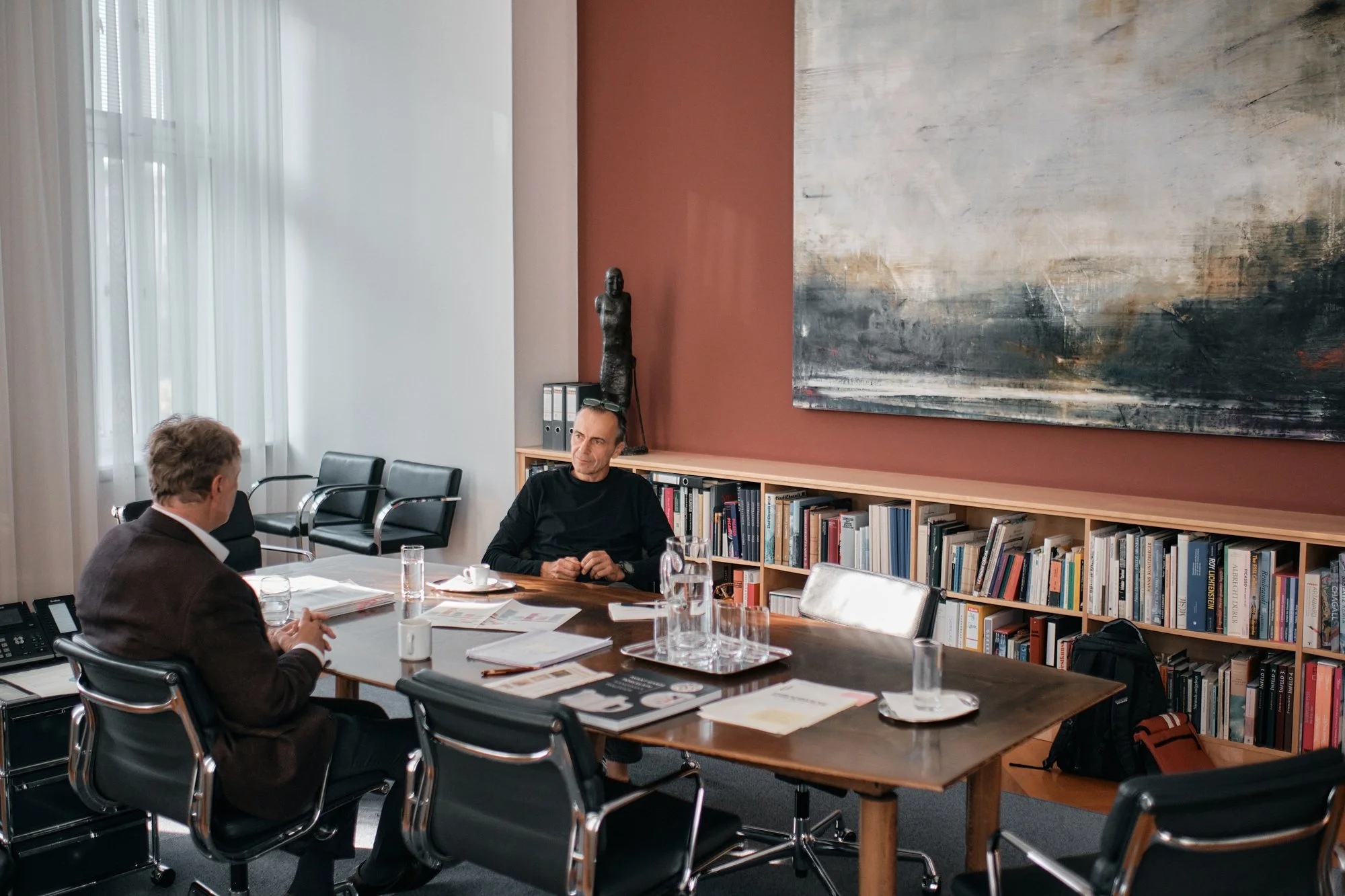 Drei Personen sitzen in einem Besprechungsraum an einem Tisch mit Getränken. Im Hintergrund sind Bücherregale, eine Skulptur, ein großformatiges abstraktes Gemälde und ein Fenster mit weißen Vorhängen zu sehen.