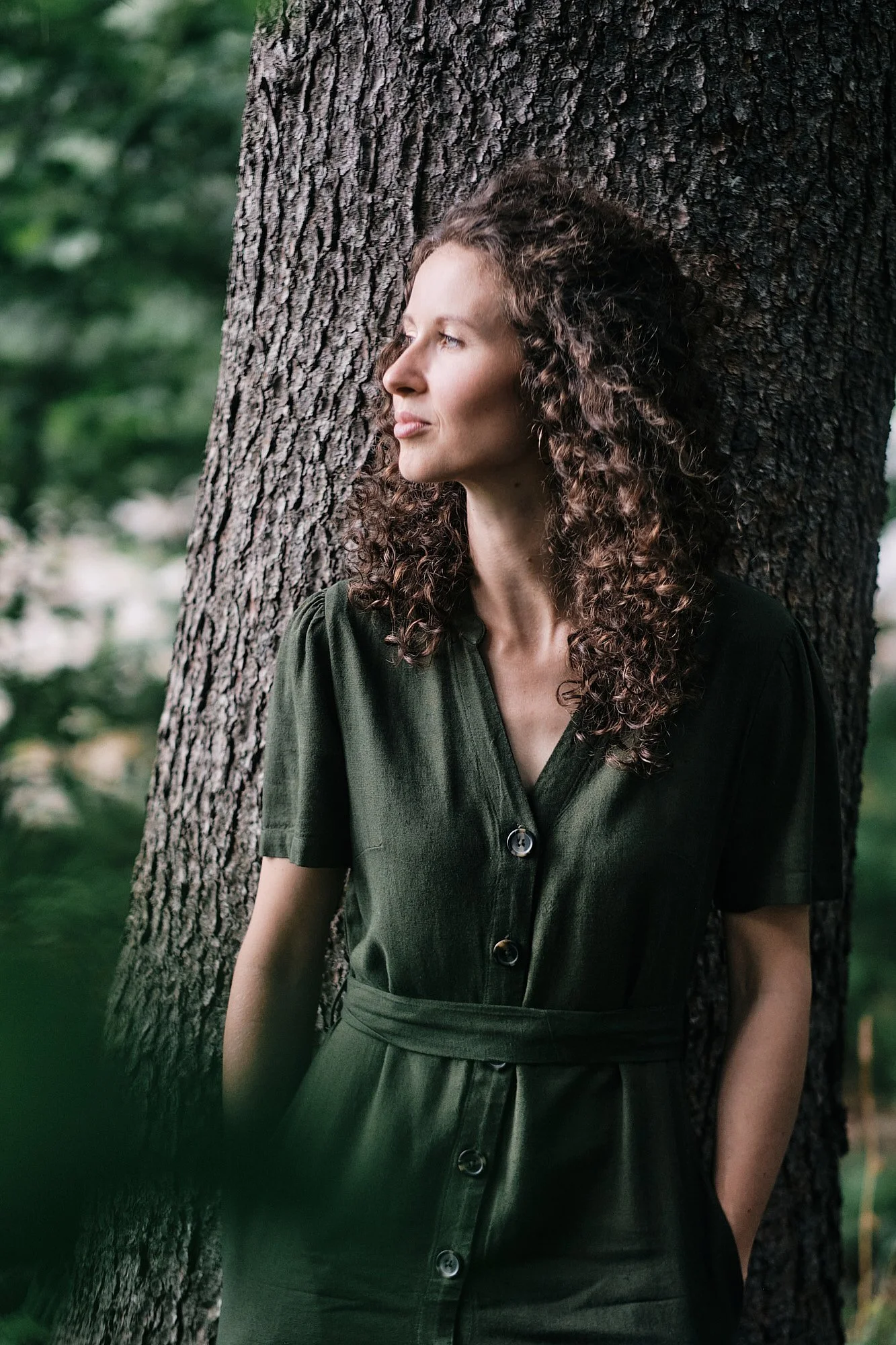 Eine Frau mit lockigem braunem Haar lehnt an einem Baum im Wald, trägt eine dunkelgrüne Bluse mit Knöpfen und einen dunkelgrünen Rock, schaut nach links, umgeben von grünen Blättern und Bäumen.