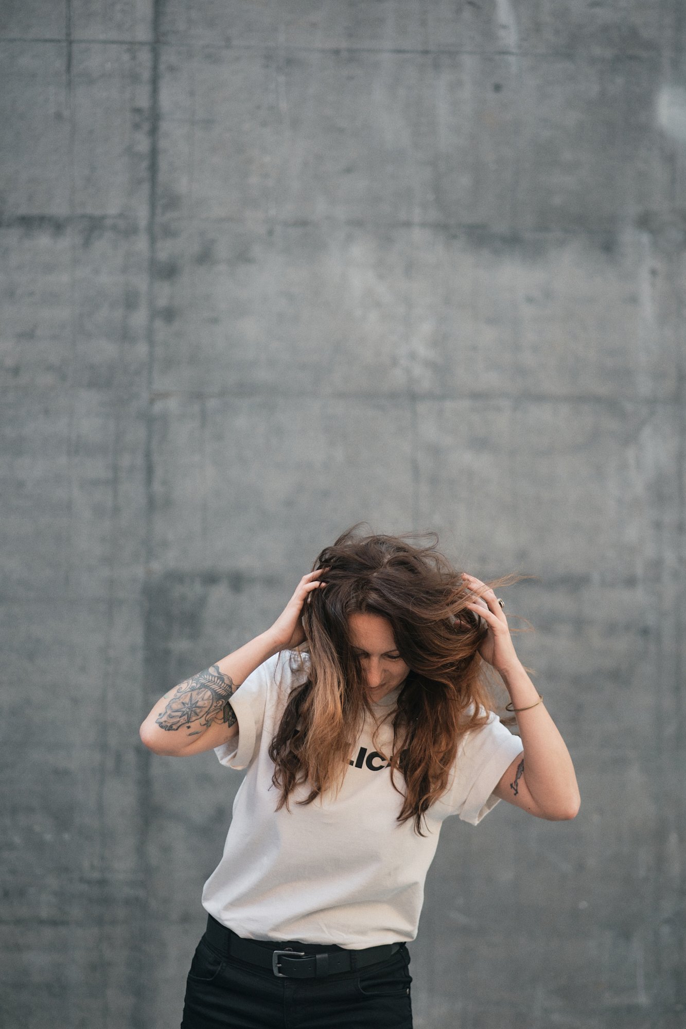 Junge Frau mit langem braunem Haar, tattoo am linken Arm, trägt ein weißes T-Shirt, steht vor einer grauen Wand, hält sich am Kopf und schaut nach unten.
