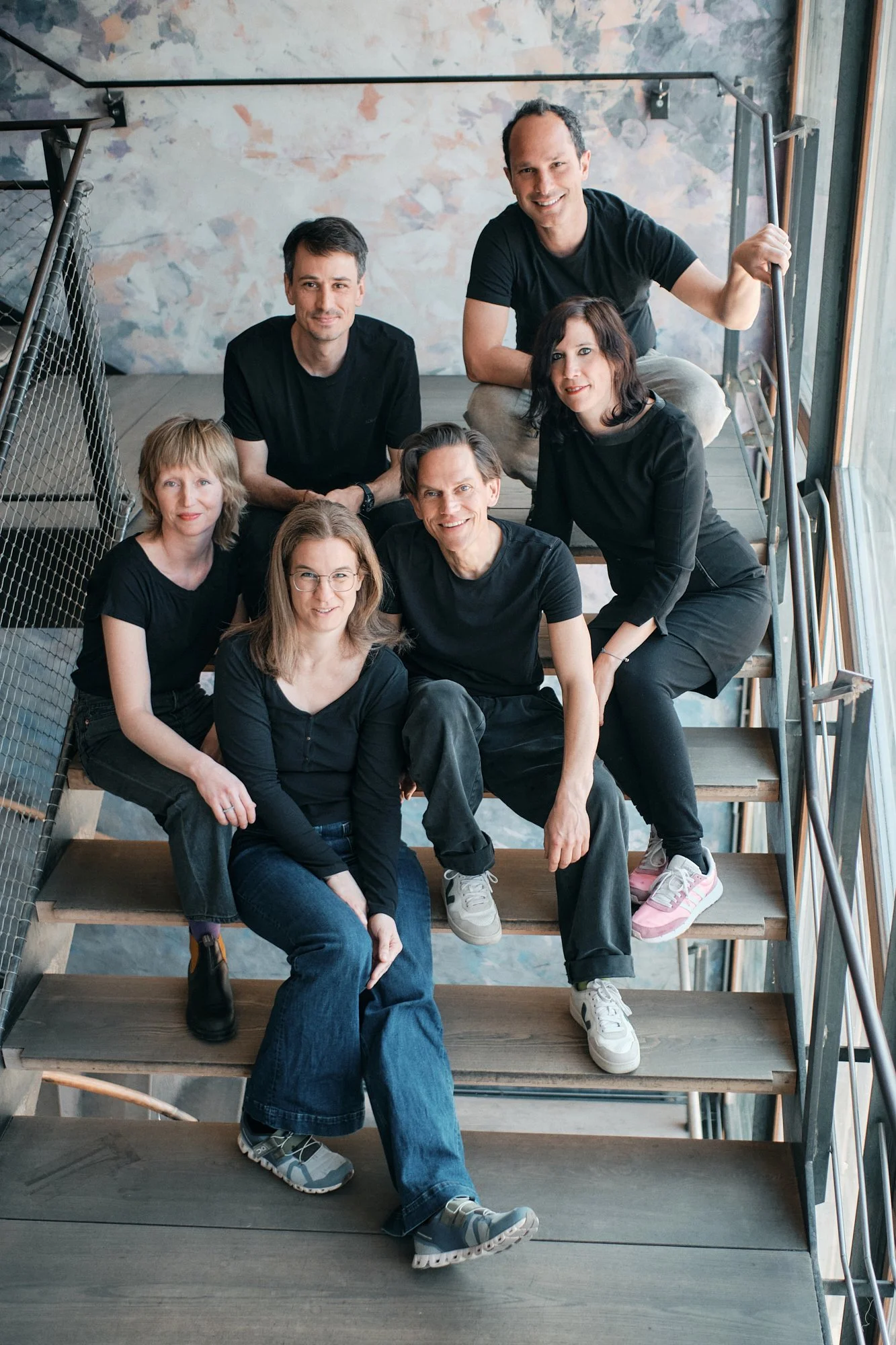 Gruppenzusammenkunft auf einer Treppe in einem modernen Raum mit Wandmalerei im Hintergrund, sechs Personen lächelnd.