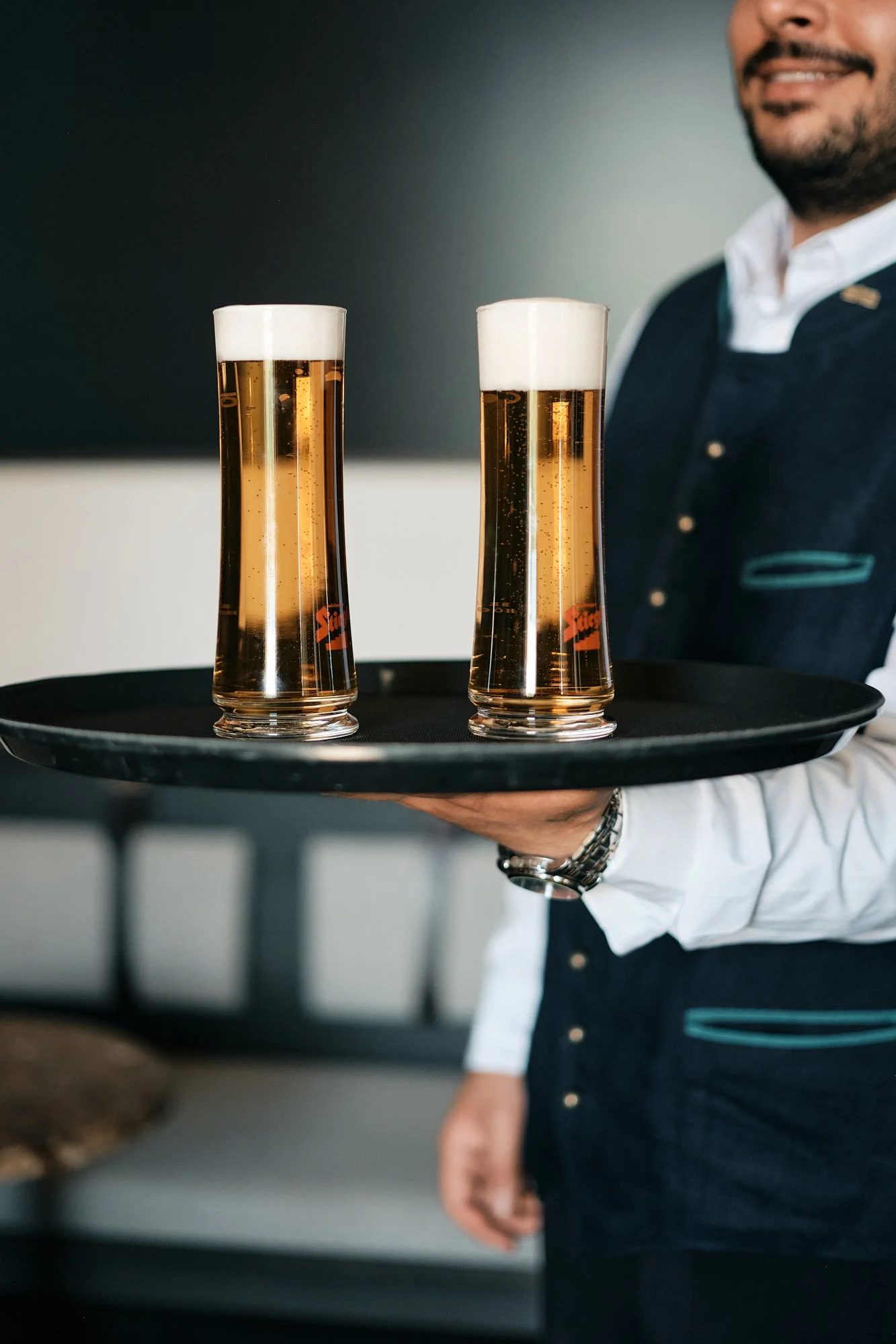Ein Kellner trägt einen runden schwarzen Tablett mit zwei hohen Biergläsern mit hellem Bier und Schaum.