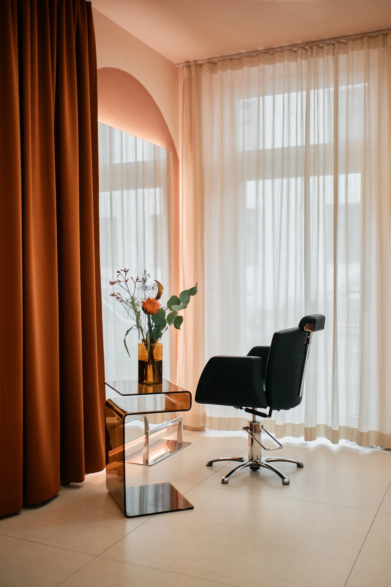 Ein stilvoll eingerichteter Raum mit großen Fenstern, hellen Vorhängen, einem schwarzen Bürostuhl, einem kleinen runden Beistelltisch mit einer Vase voller Blumen, welche auf einem transparenten Couchtisch stehen.